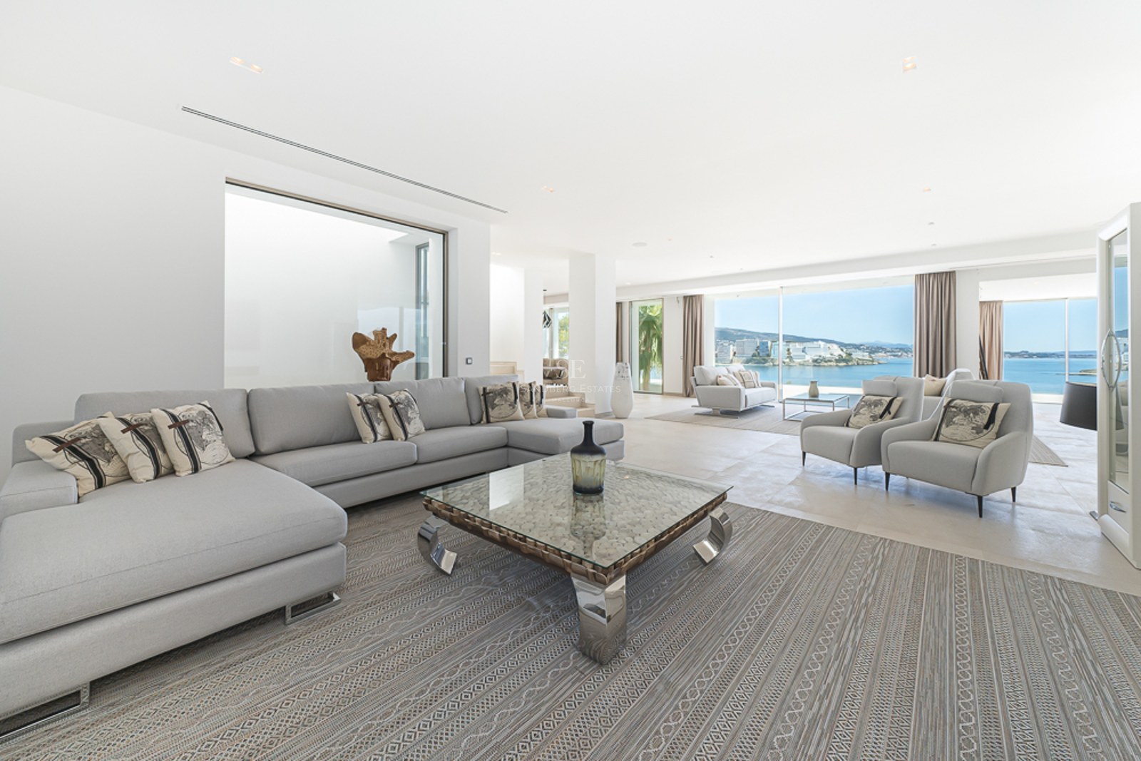 Amplio salón moderno con sofás gris claro, mesa de centro de cristal y grandes ventanales con vistas al mar Mediterráneo.