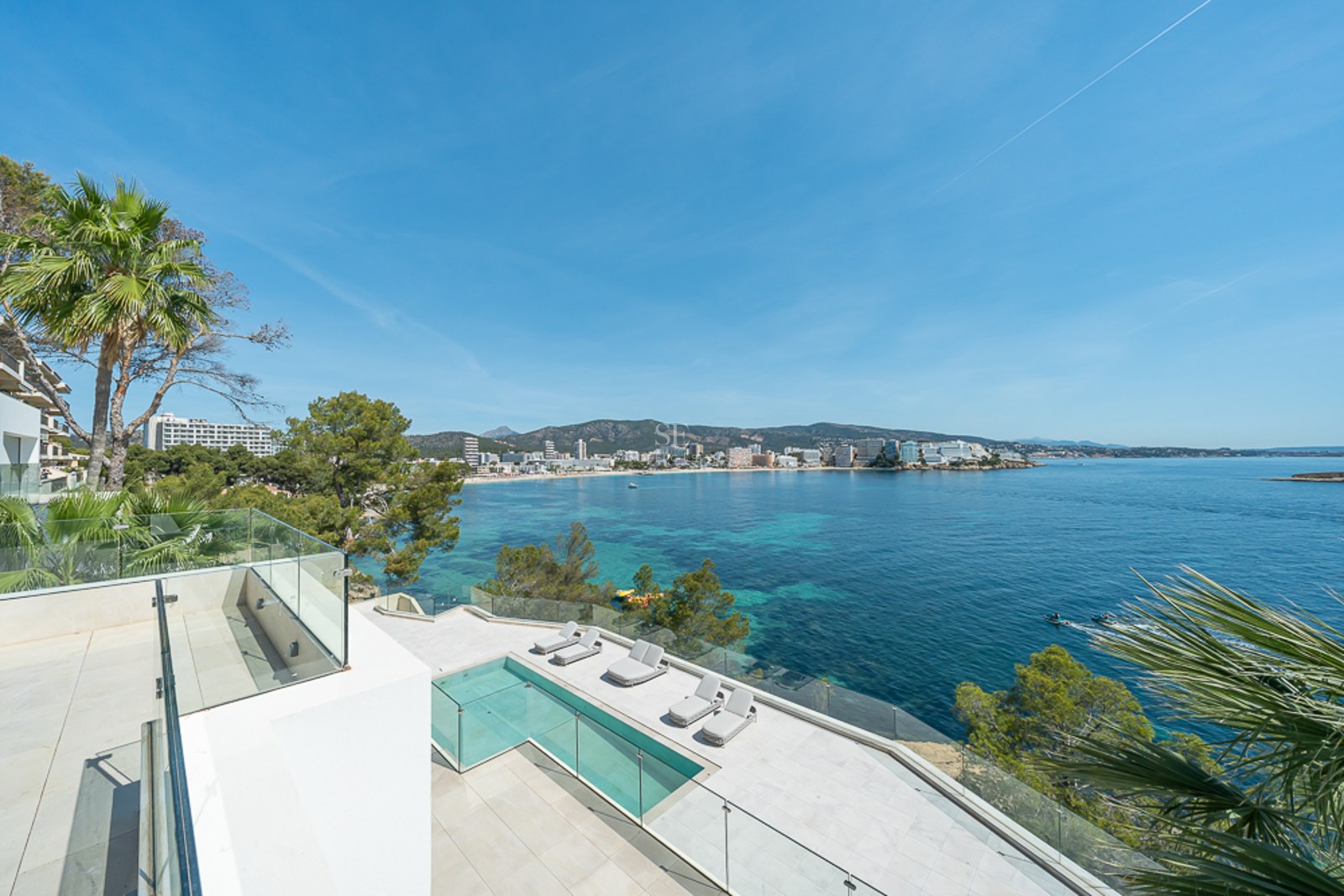 Terraza blanca moderna con piscina infinita y barandillas de cristal con vistas a una bahía de aguas turquesas.