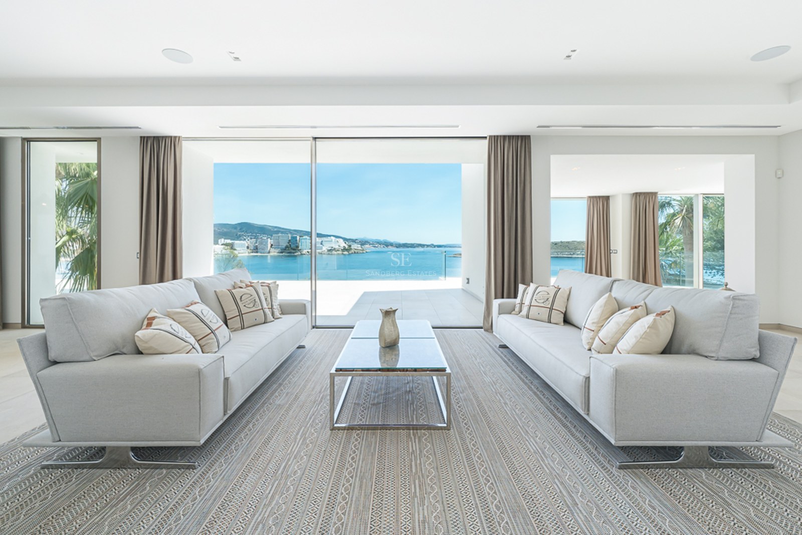 Salón luminoso con dos sofás grises, mesa de centro de cristal y grandes ventanales con vistas al mar.