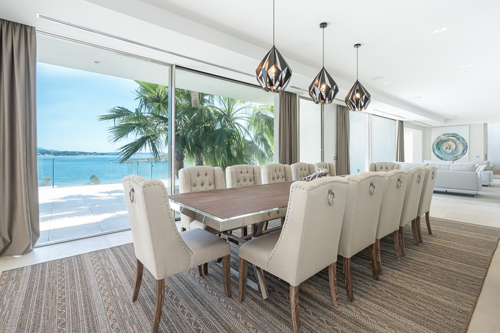 Elegante comedor con mesa larga, sillas tapizadas de color beige y grandes ventanales con vistas al mar y palmeras.