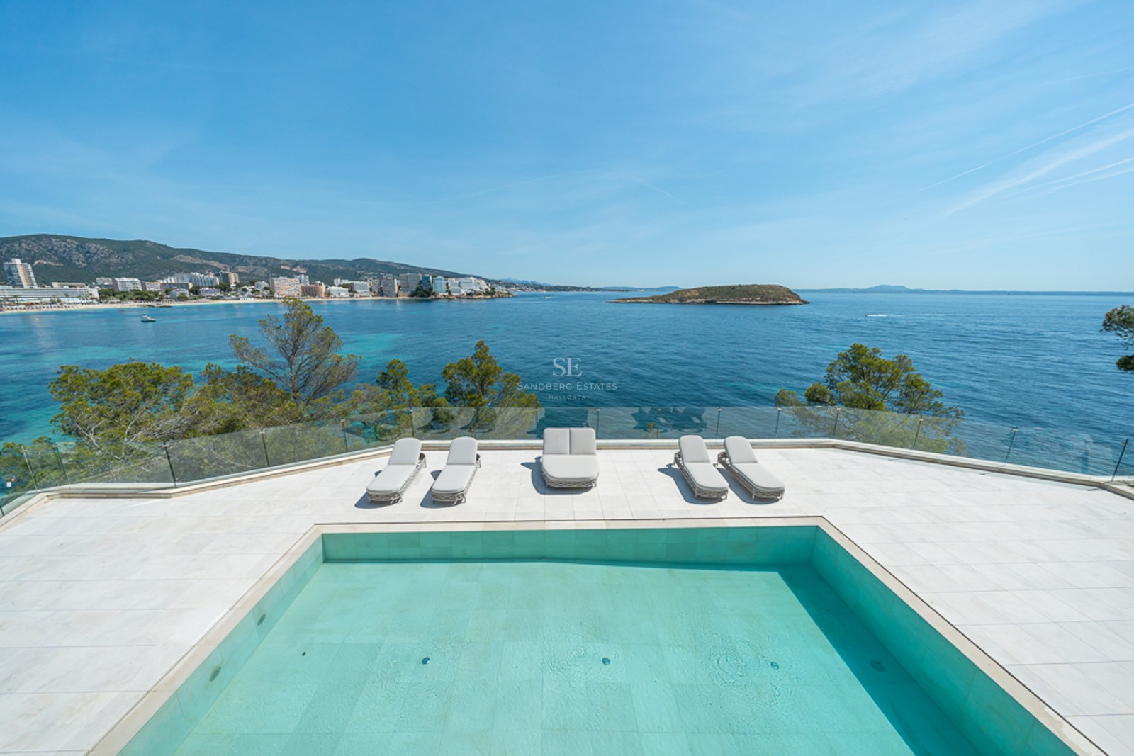 Piscina moderna y tumbonas en una terraza blanca con vistas a una bahía turquesa y la costa.