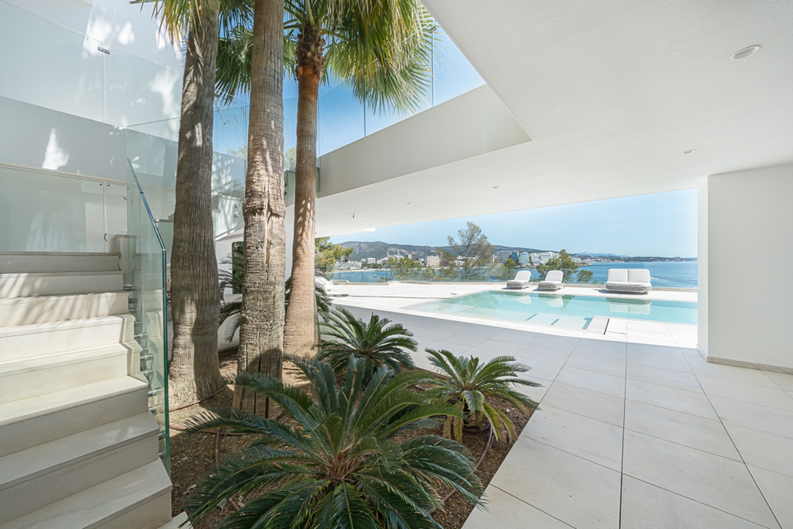 Terraza de villa moderna con piscina, palmeras y barandillas de cristal con vistas al mar Mediterráneo azul.