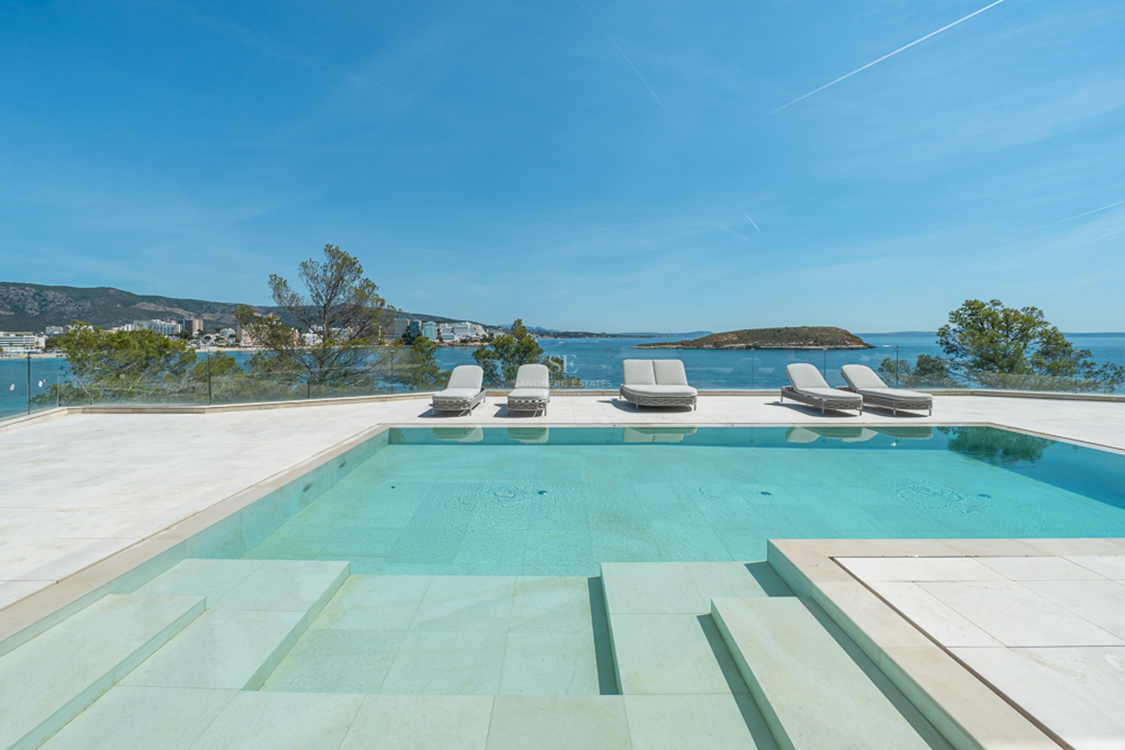 Una piscina turquesa en una terraza de piedra con tumbonas con vistas al mar Mediterráneo y una isla lejana.