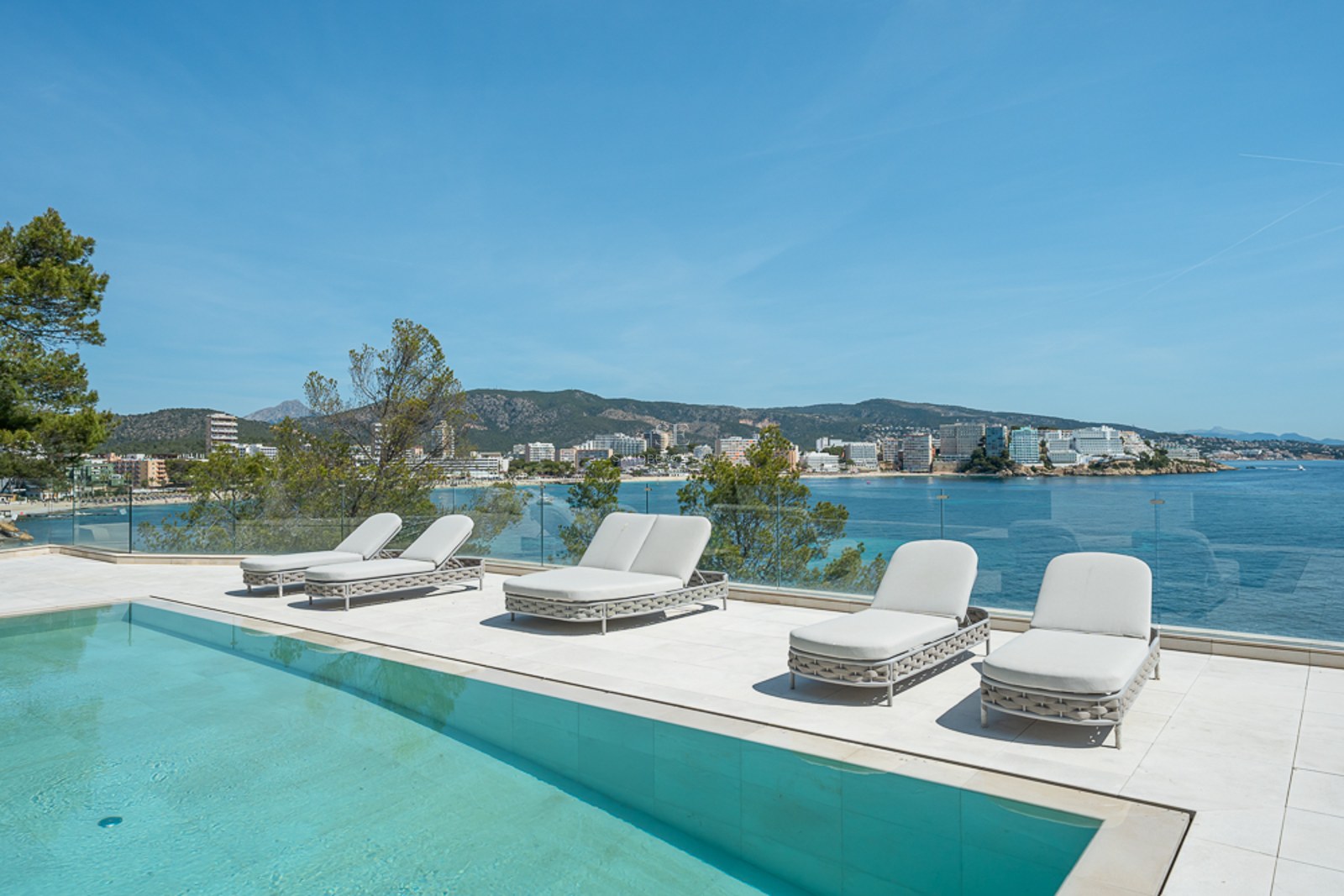 Terraza blanca moderna con piscina turquesa, tumbonas y barandilla de cristal con vistas a una bahía mediterránea.