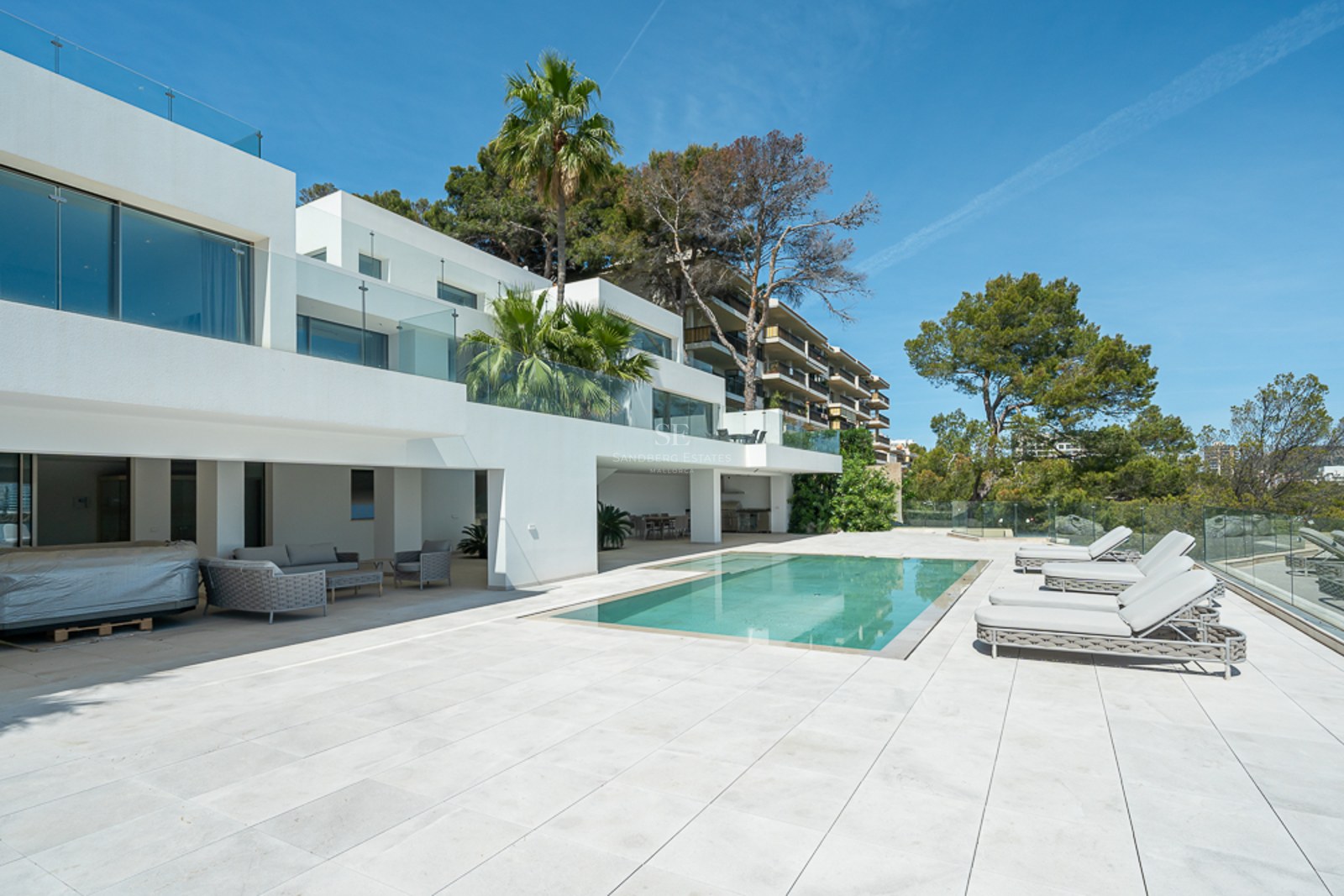 Villa blanca moderna con una piscina rectangular, tumbonas grises y barandillas de cristal bajo un cielo azul.