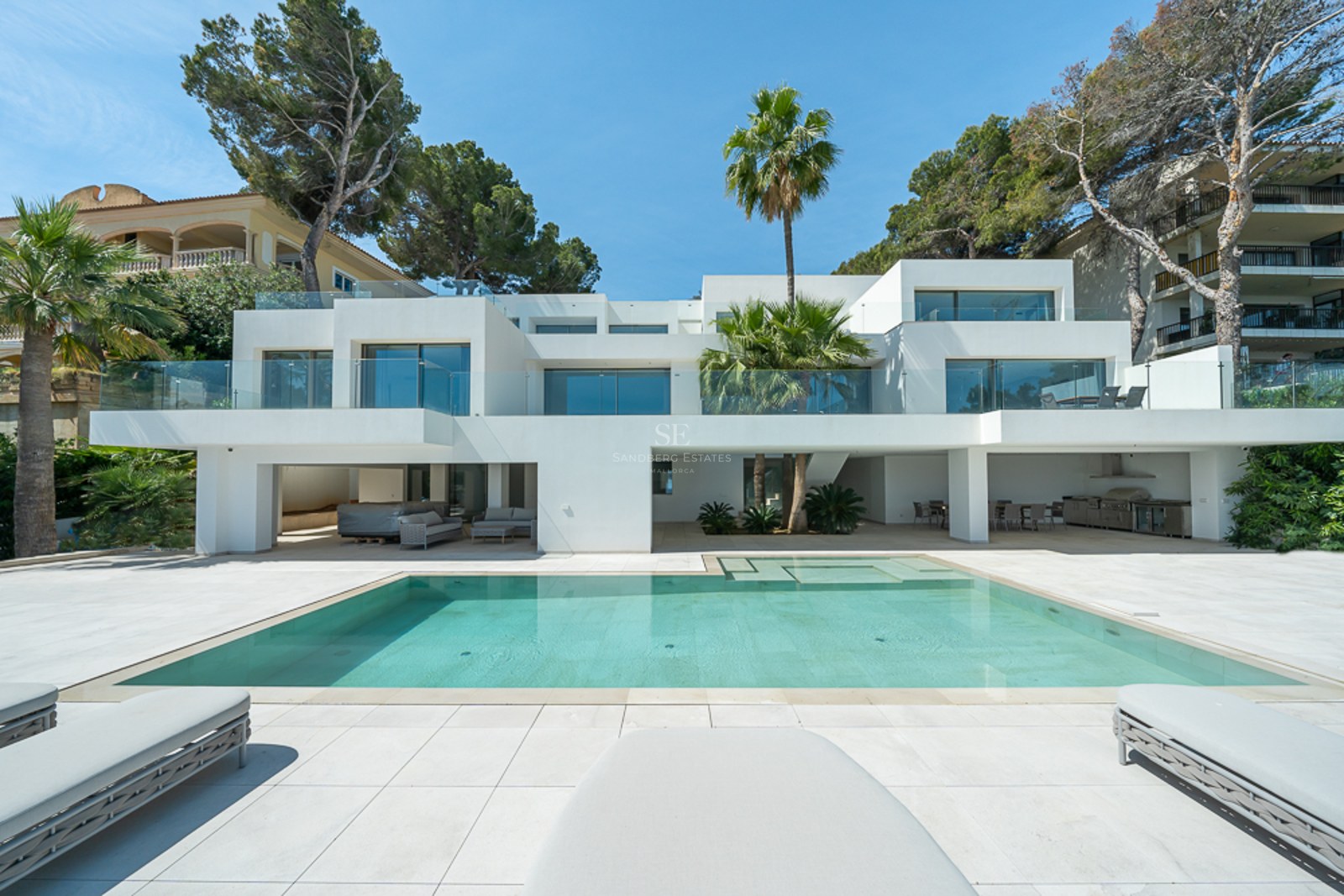 Villa moderna blanca con una gran piscina turquesa, barandillas de cristal y palmeras bajo un cielo despejado.