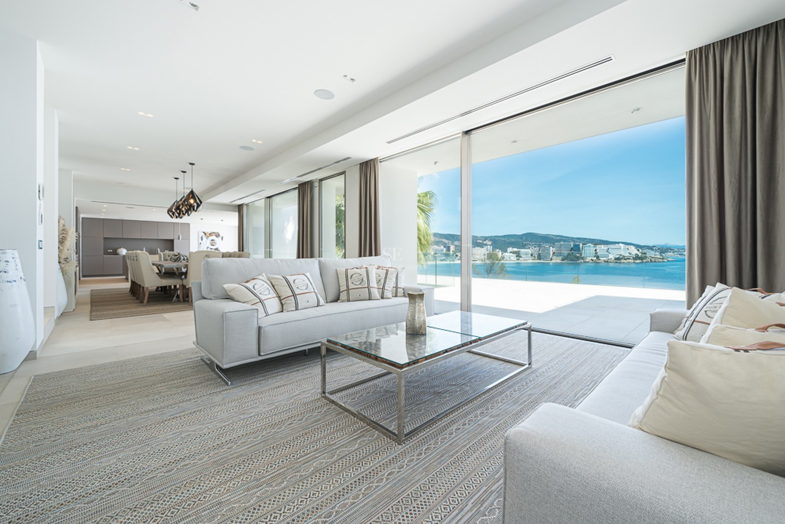 Salón moderno con sofás grises y grandes ventanales que muestran una vista panorámica del mar y la costa.