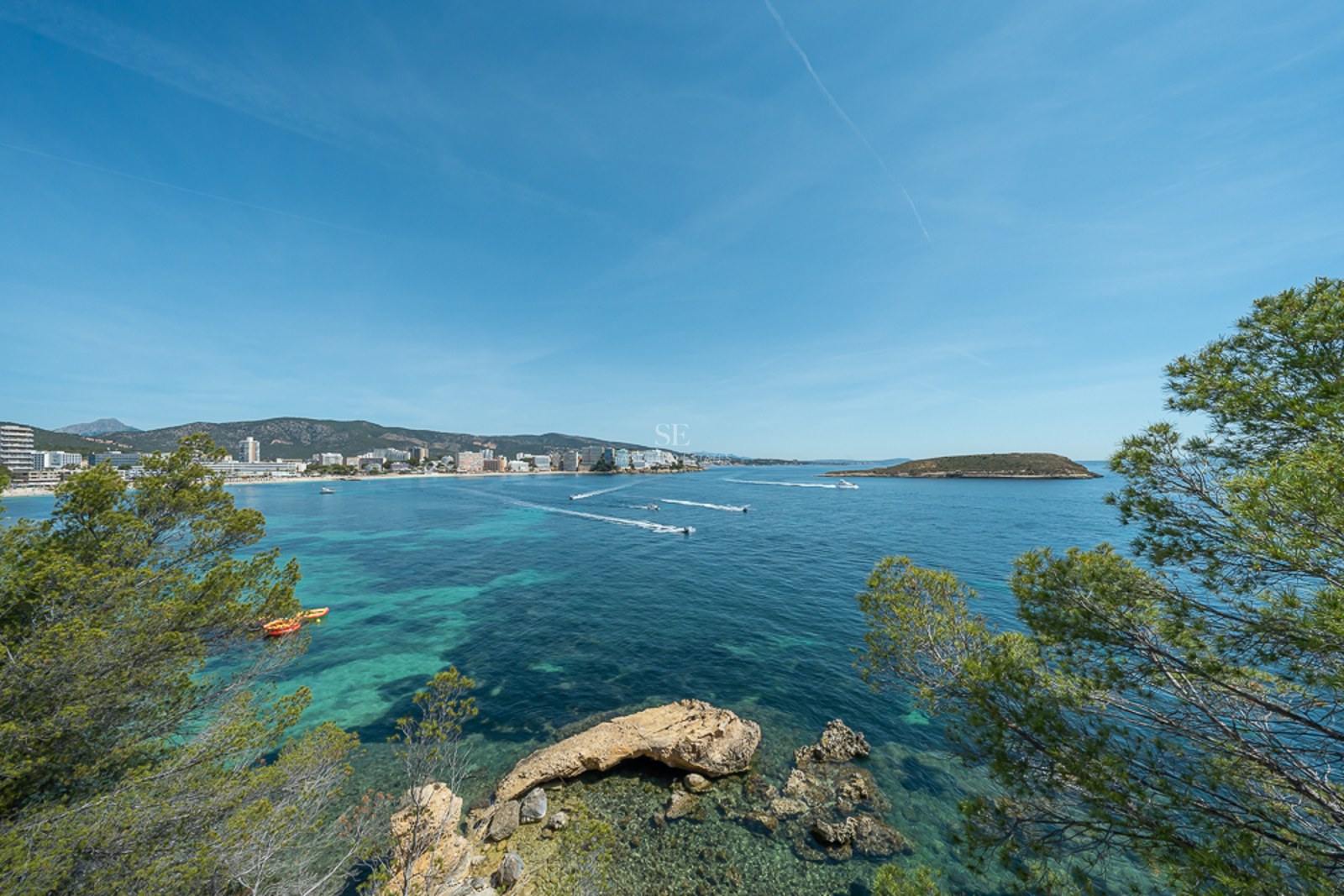 Vista de una bahía de aguas turquesas con barcos, rodeada de pinos y una ciudad costera al fondo.