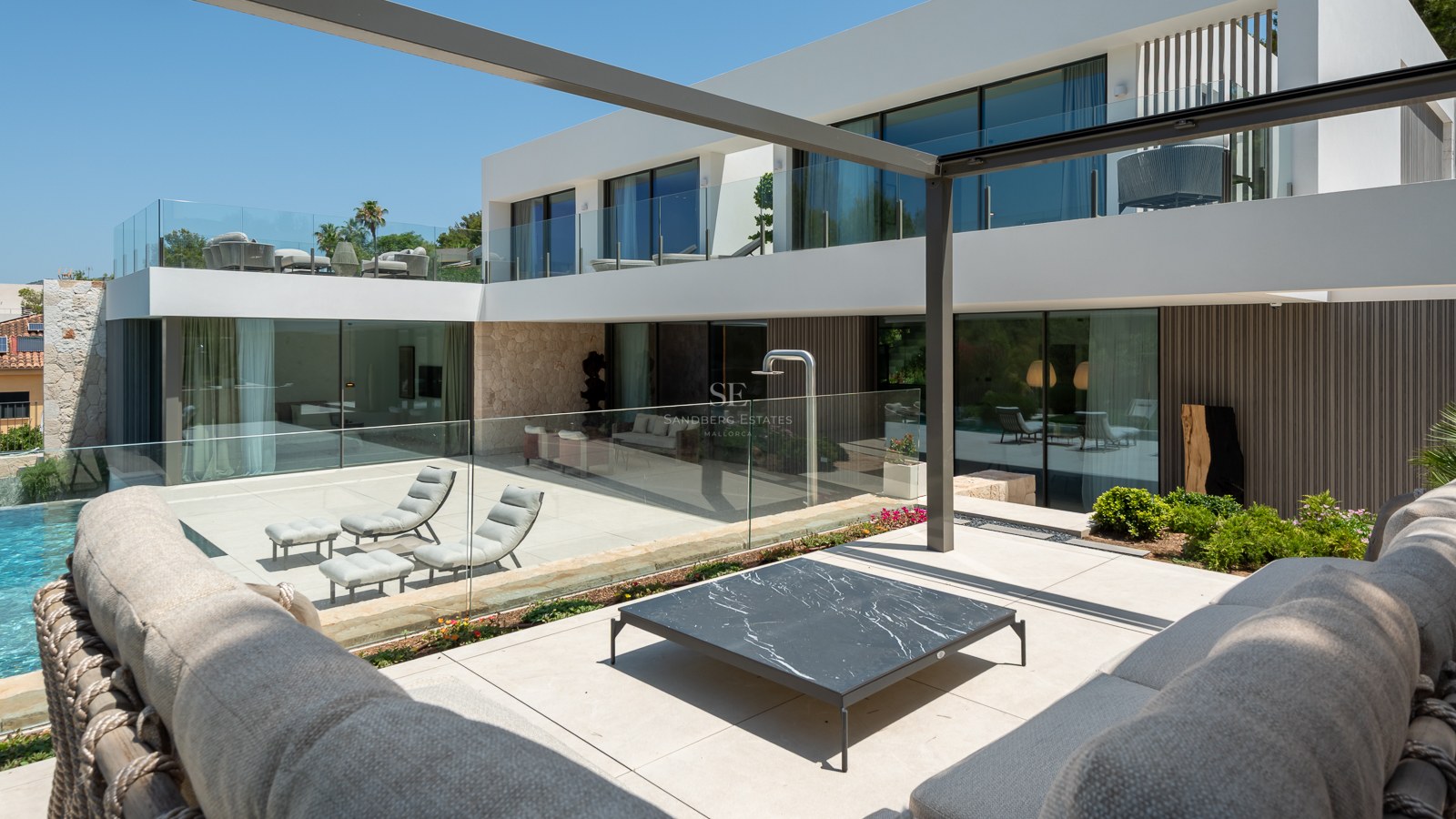 Villa blanche moderne avec balustrades en verre, murs en pierre et terrasse ensoleillée avec mobilier de salon design.
