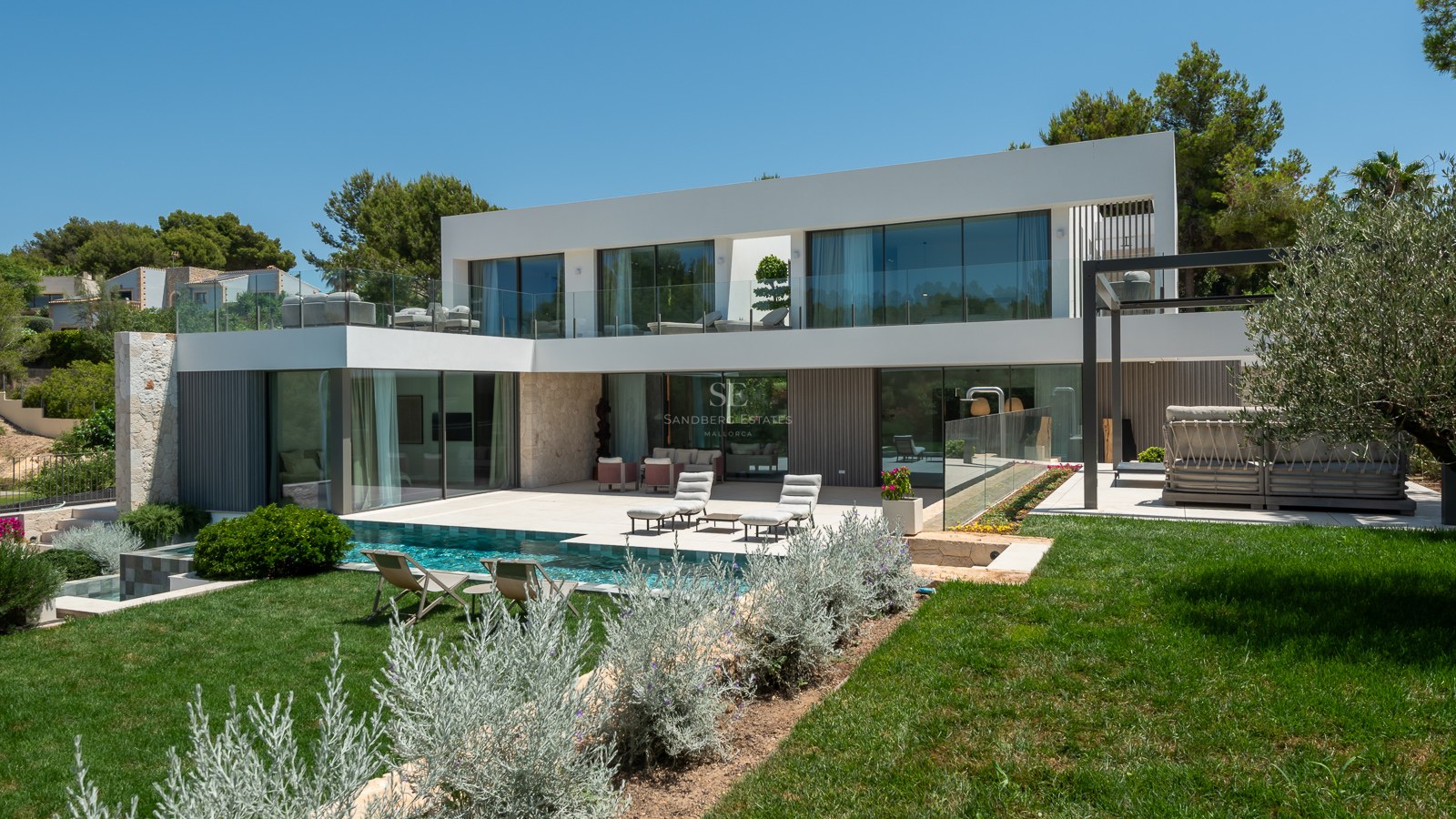 Façade arrière d'une villa moderne blanche avec piscine turquoise, pelouse, accents en pierre et balcons en verre.