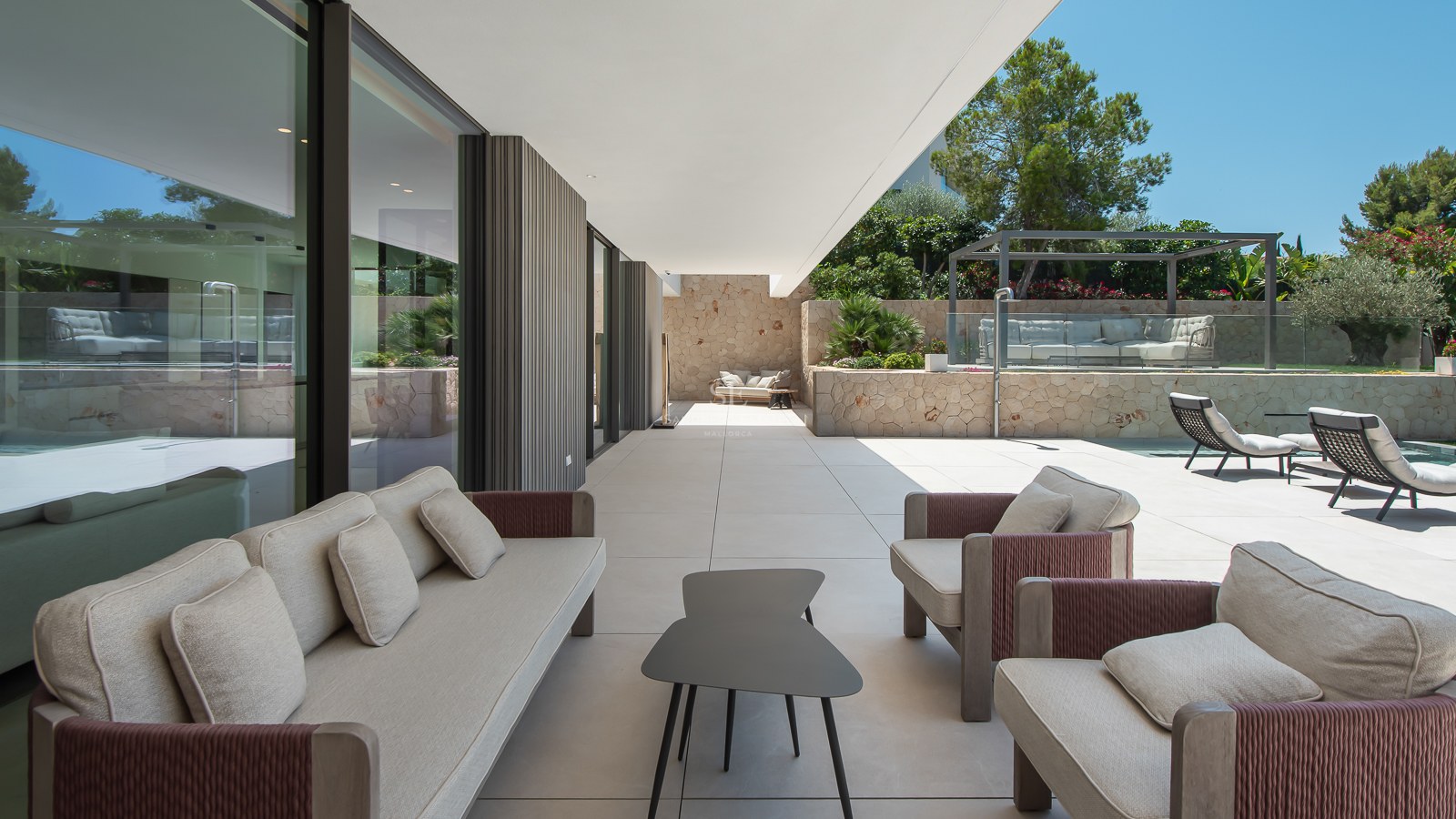 Terrasse couverte avec mobilier gris, murs en pierre naturelle et piscine sous un ciel bleu.