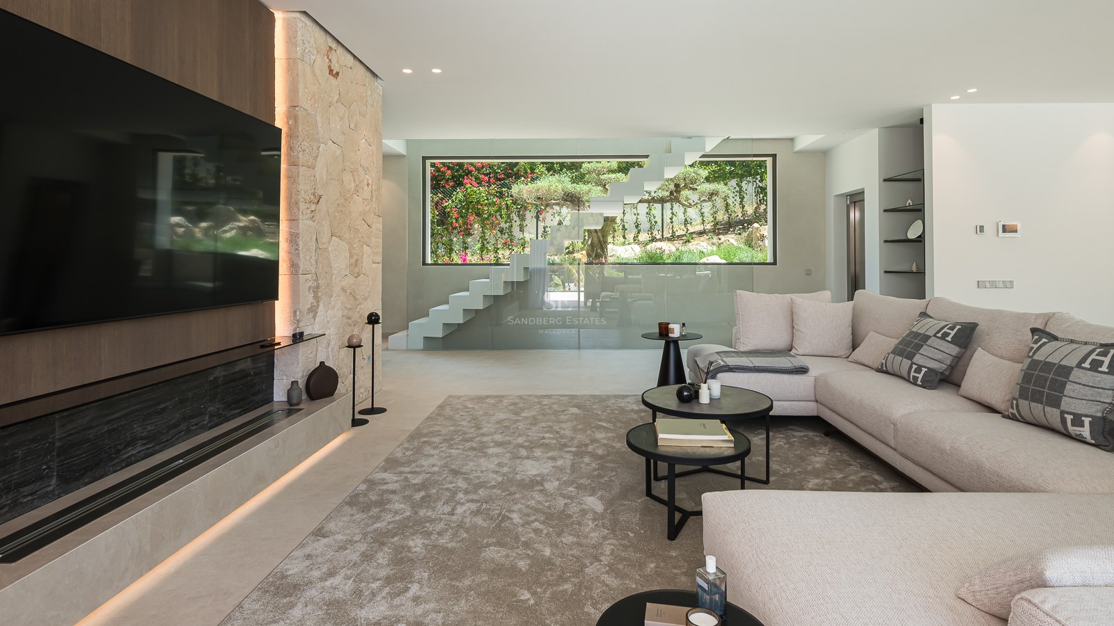 Salon moderne avec canapé beige, mur en pierre avec cheminée intégrée et escalier en verre vue sur jardin.