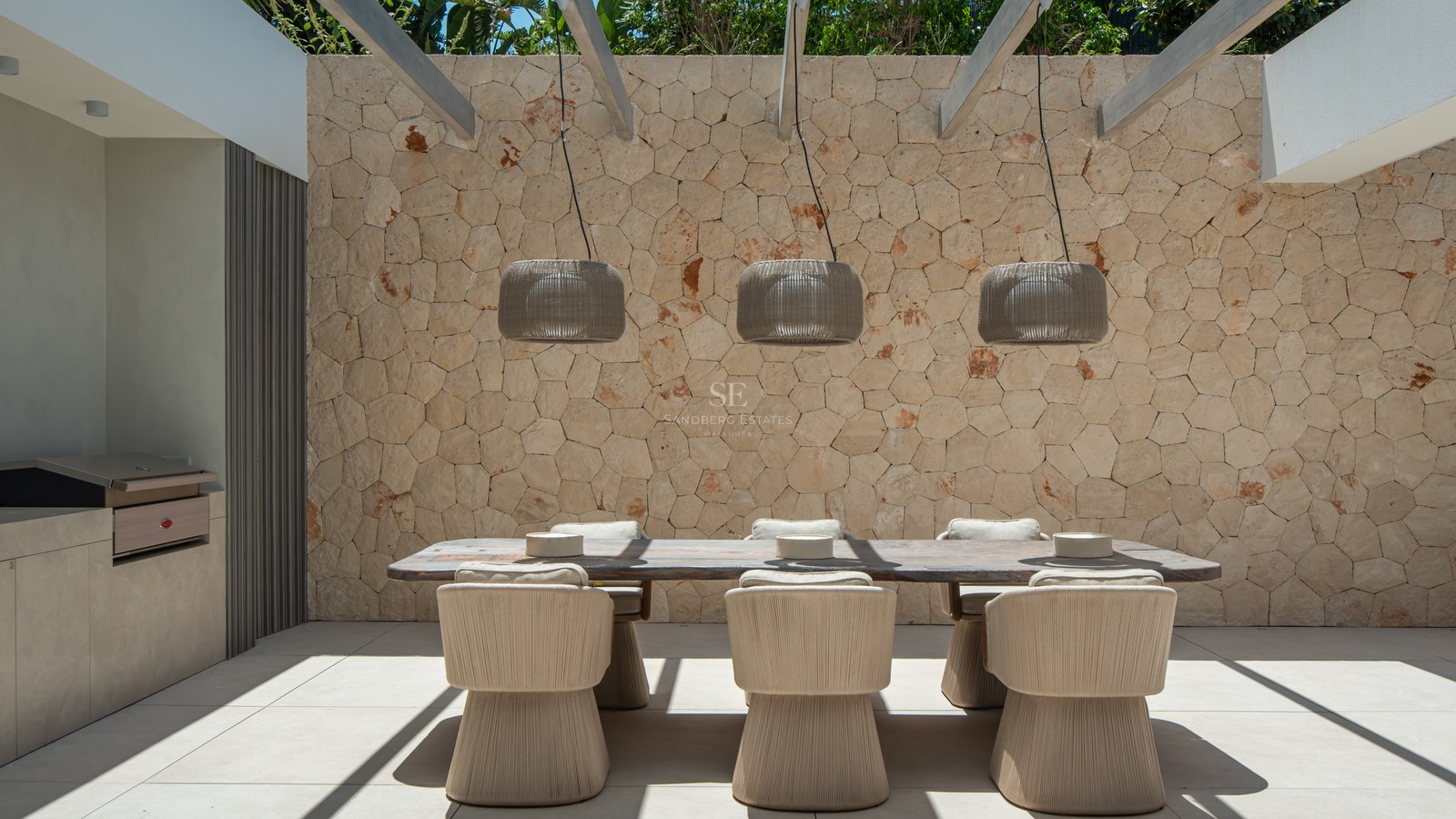 Espace repas extérieur chic avec table en bois, chaises en corde et pergola devant un mur en pierres apparentes.