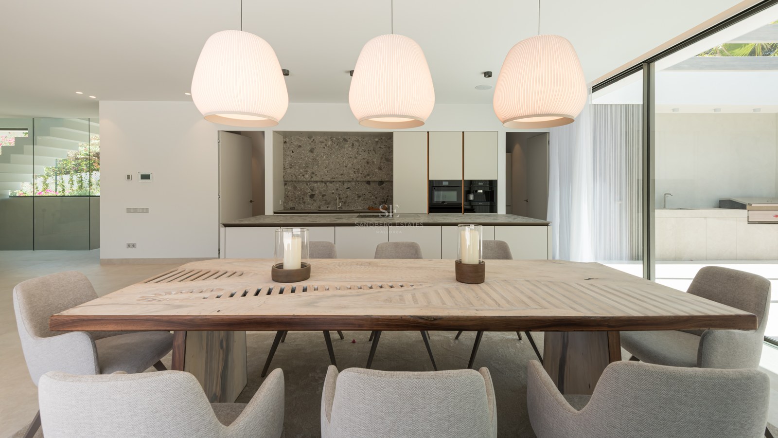 Salle à manger moderne avec table en bois massif, chaises grises et cuisine élégante avec îlot en pierre en arrière-plan.