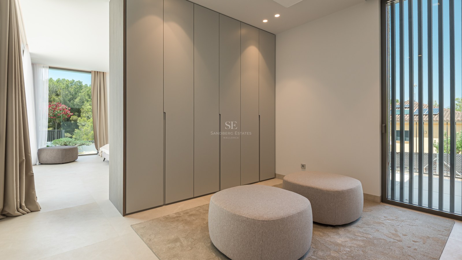 Dressing minimaliste avec armoires du sol au plafond, deux poufs en tissu et grandes fenêtres avec persiennes.