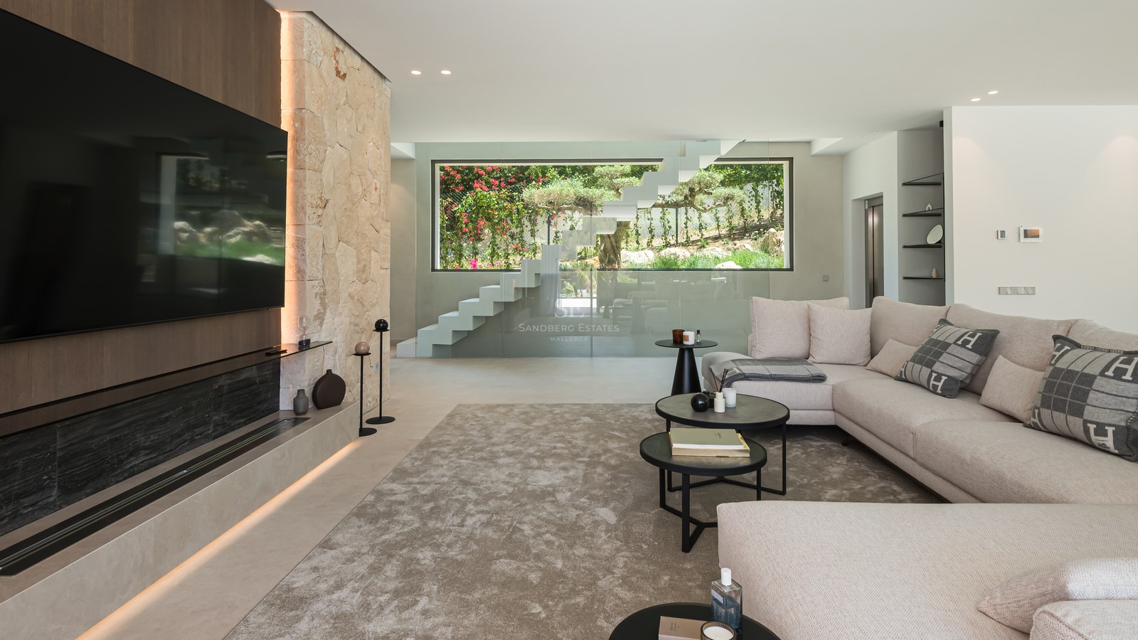 Salon moderne avec grand canapé, mur en pierre naturelle, TV intégrée et escalier suspendu en verre.