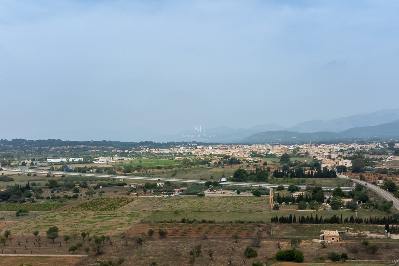 Weite Landschaft einer mallorquinischen Stadt, umgeben von Feldern und Bergen unter dunstigem blauem Himmel.