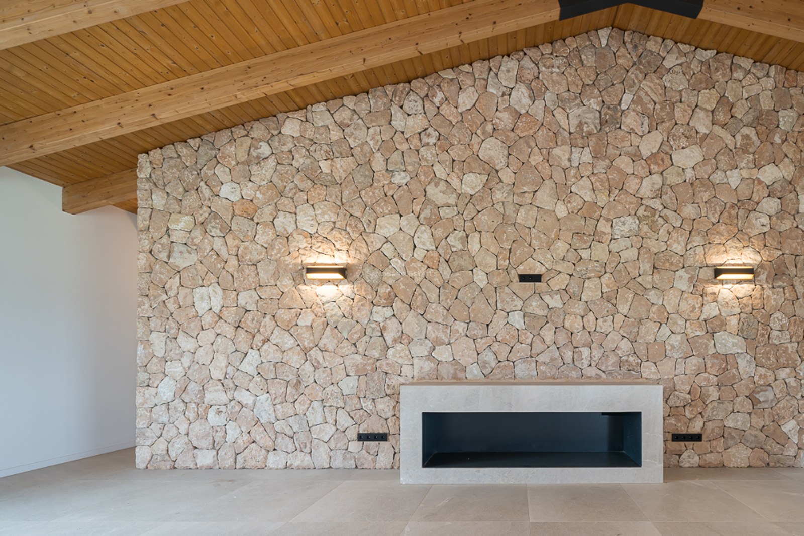Innenansicht mit großer Natursteinwand, modernem Kamin und hoher Holzdecke mit Balken.