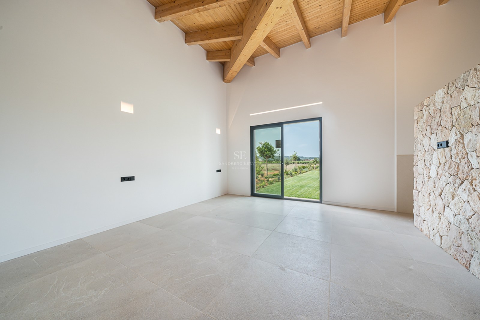 Leerer moderner Raum mit hoher Holzbalkendecke, Natursteinwand und Glastür zum grünen Garten.