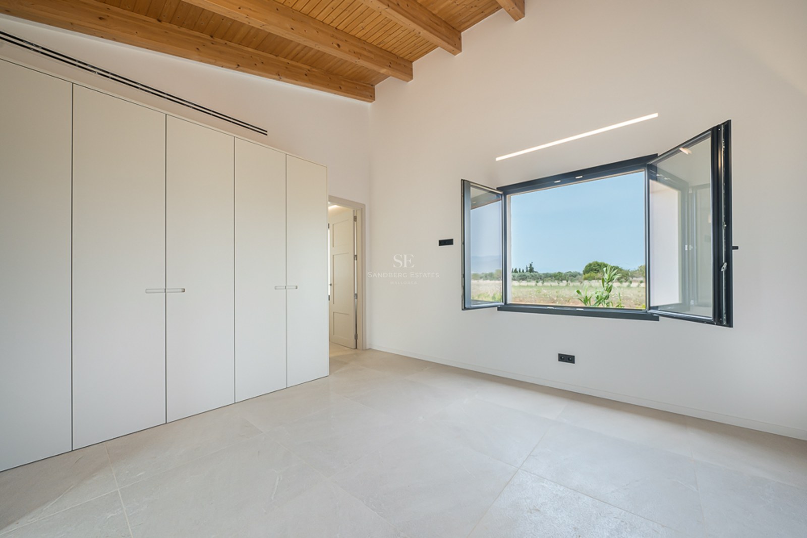 Helles Schlafzimmer mit hohen Holzbalkendecken, weißen Einbauschränken und offenem Fenster mit Naturblick.