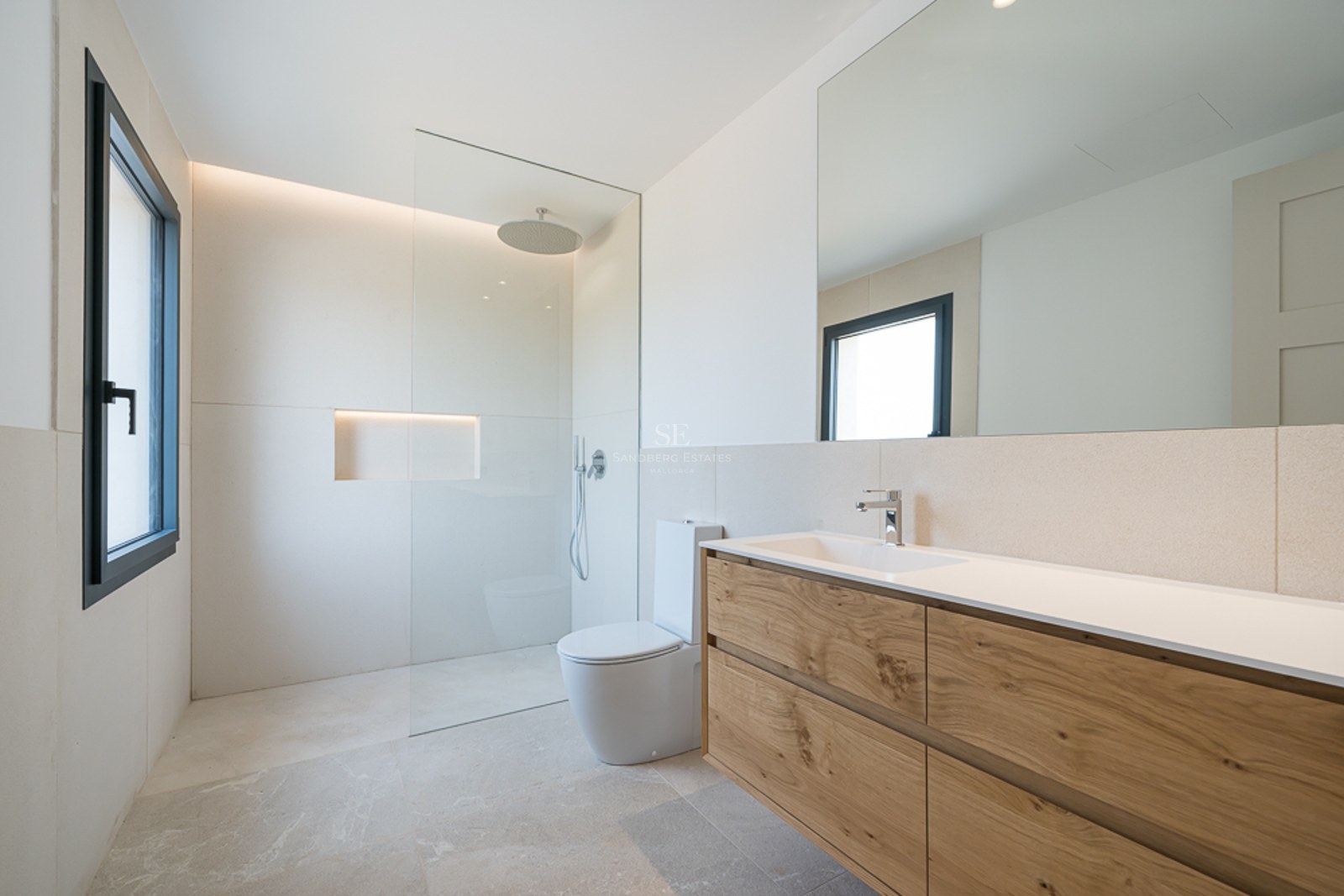 Helles Badezimmer mit Holzwaschtisch, großem Spiegel, bodengleicher Dusche und beigen Natursteinfliesen.