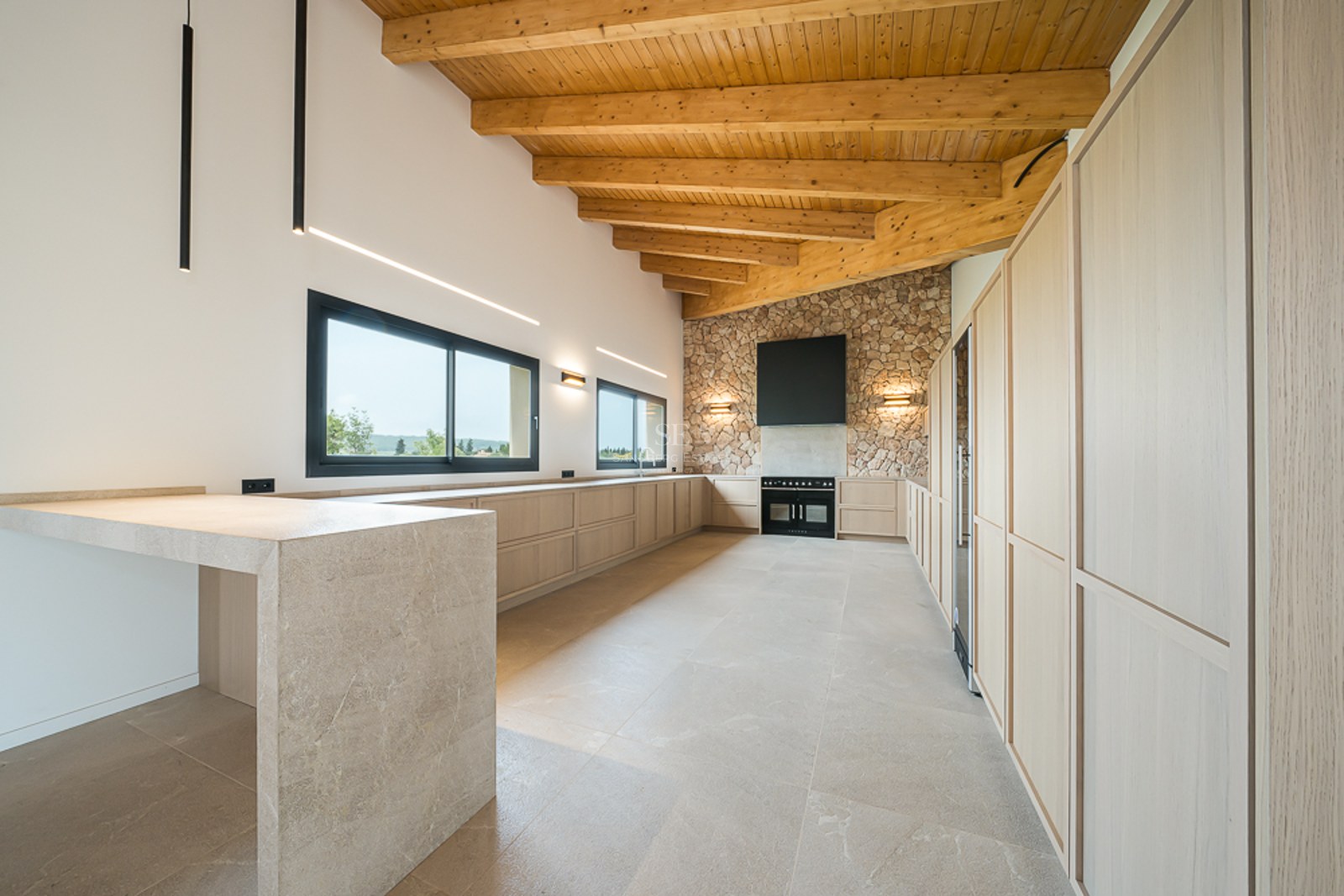 Geräumige moderne Küche mit hohen Holzbalkendecken, Natursteinwand, beigen Schränken und schwarzen Geräten.