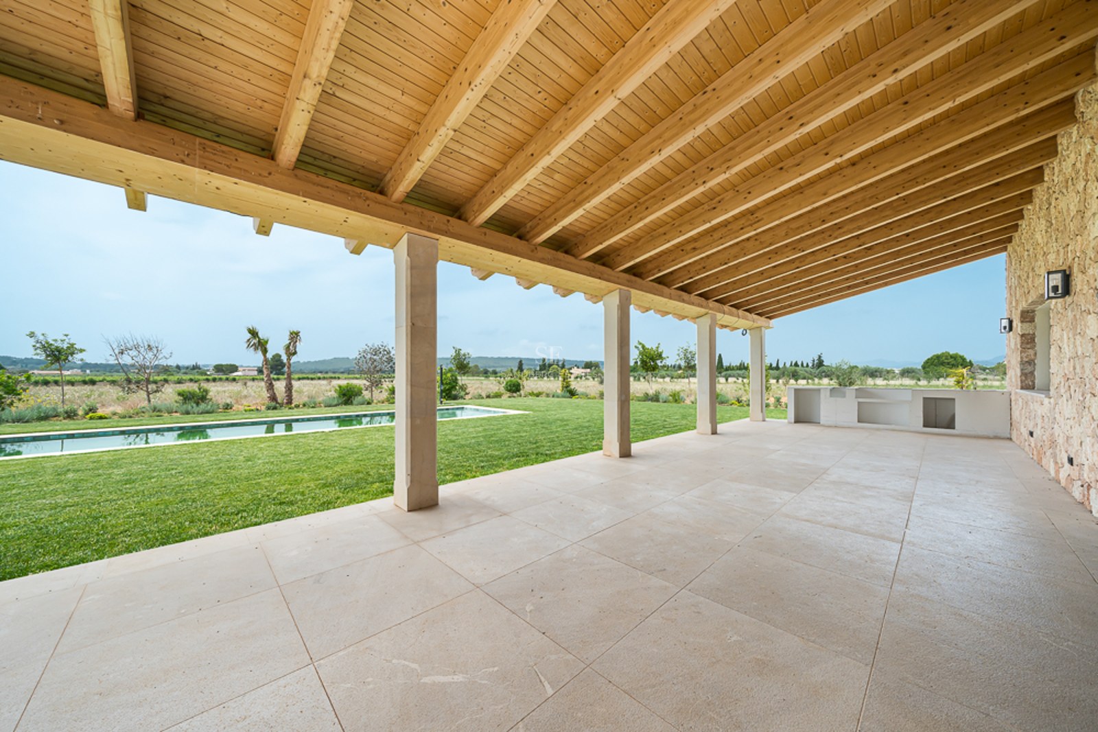 Weite überdachte Terrasse mit Natursteinboden und Holzbalkendecke mit Blick auf Garten und Pool.