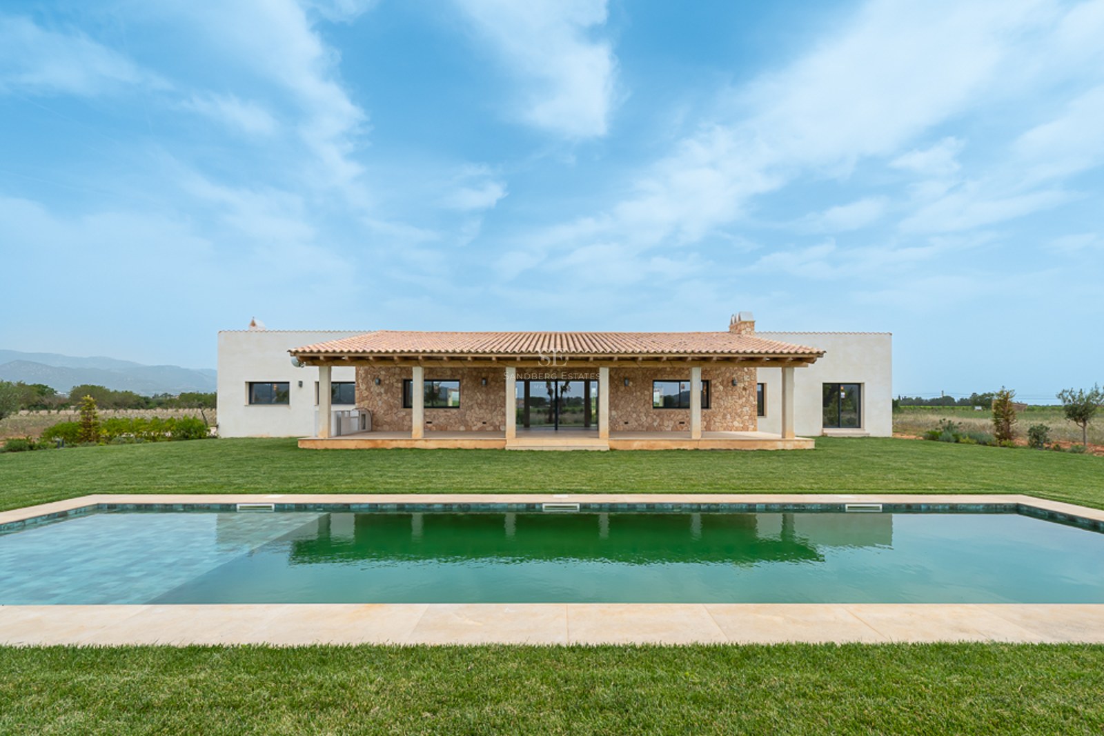 Luxusvilla mit langem rechteckigem Pool, grünem Rasen, Steinfassade und Terrakotta-Veranda unter blauem Himmel.