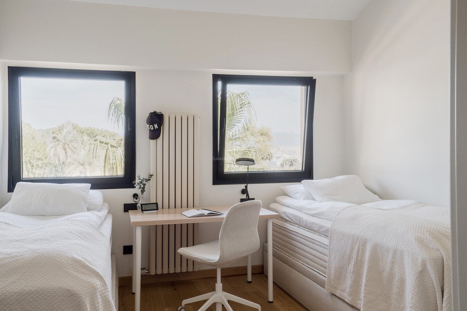 Dormitorio con dos camas individuales blancas, un escritorio central y dos ventanas con vistas a palmeras.