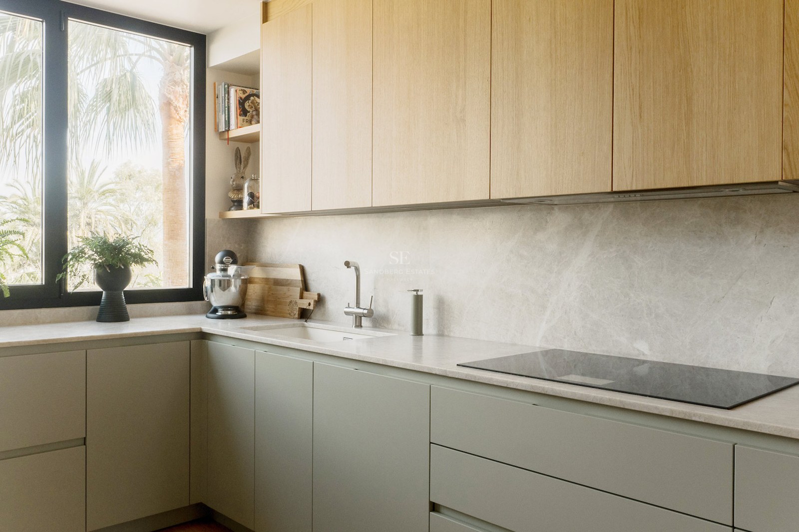 Cocina de diseño con armarios de madera clara, encimera de piedra gris y ventana con vistas a palmeras.
