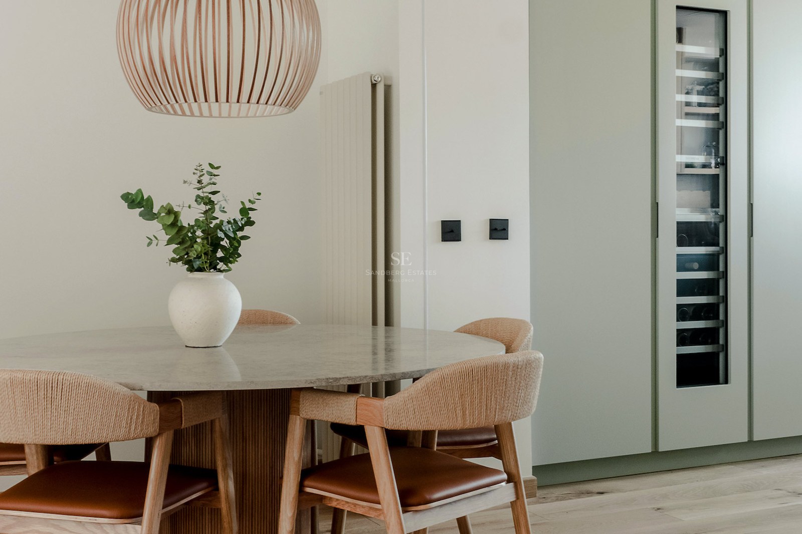 Comedor moderno con mesa de piedra, sillas de madera tejida y una vinoteca empotrada en armarios verde salvia.