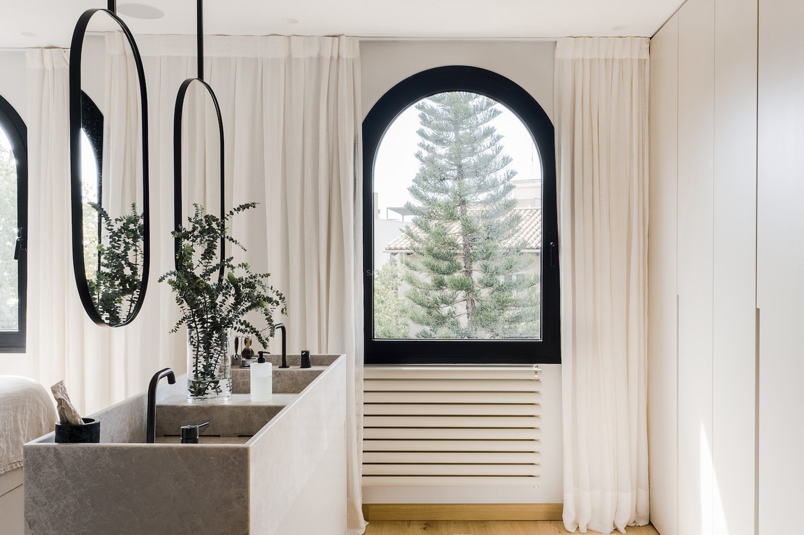 Lavabo doble de piedra con espejos ovalados negros frente a una ventana de arco con vistas a un árbol.