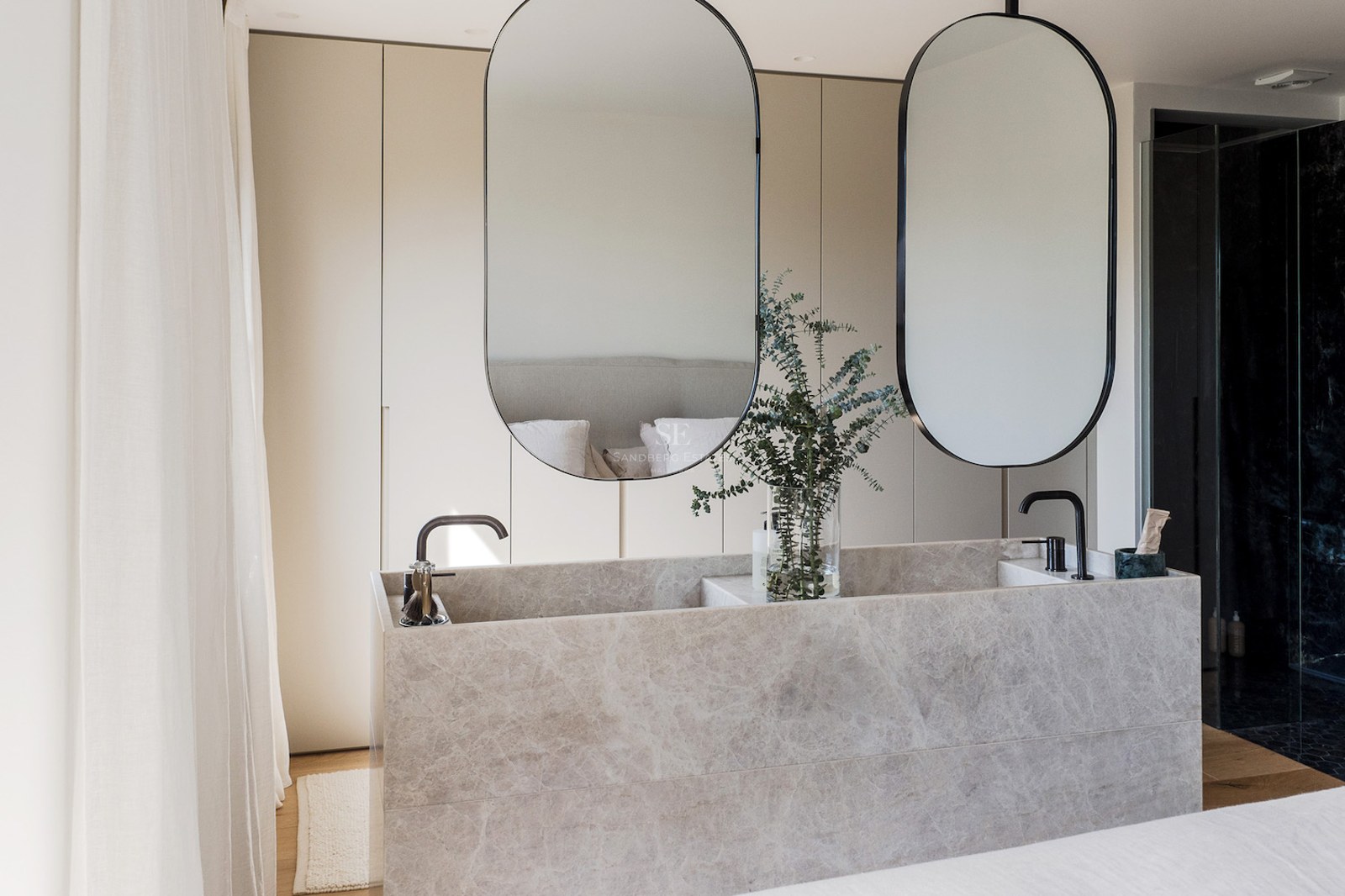 Lavabo doble moderno de piedra mármol, dos espejos ovalados y grifos negros en un baño minimalista y luminoso.