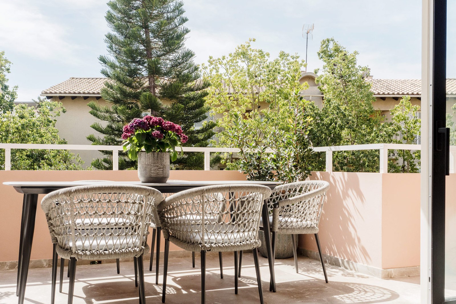 Terraza soleada con mesa negra, sillas tejidas y flores púrpuras, con vistas a árboles exuberantes.