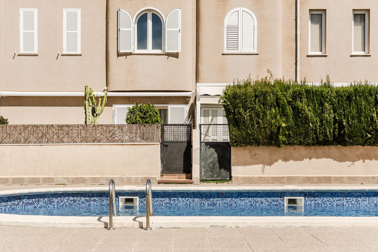 Piscina exterior con mosaicos azules frente a una fachada beige con persianas blancas.