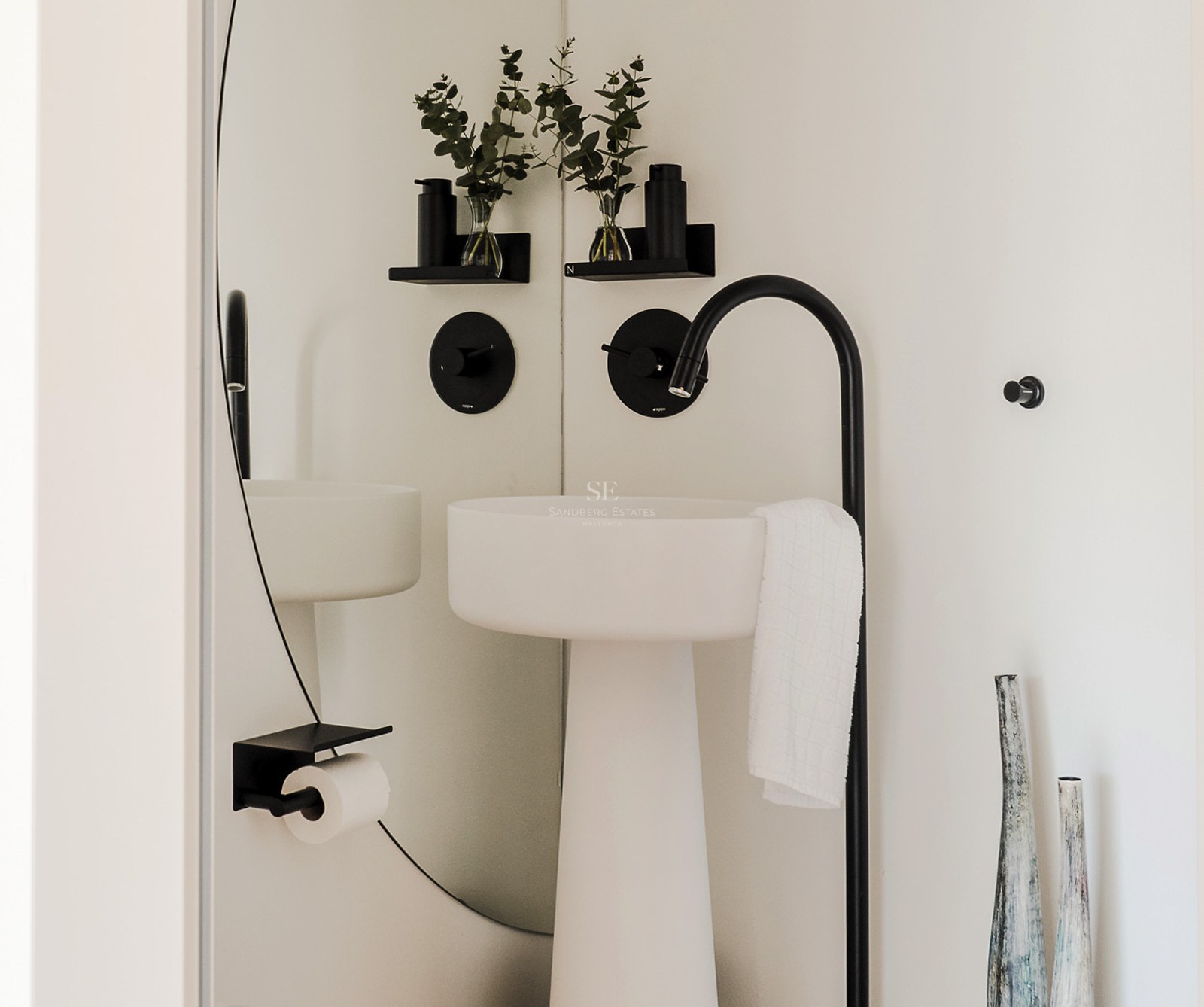 Lavabo de pedestal blanco con grifo negro mate alto, espejo redondo y ramas de eucalipto.