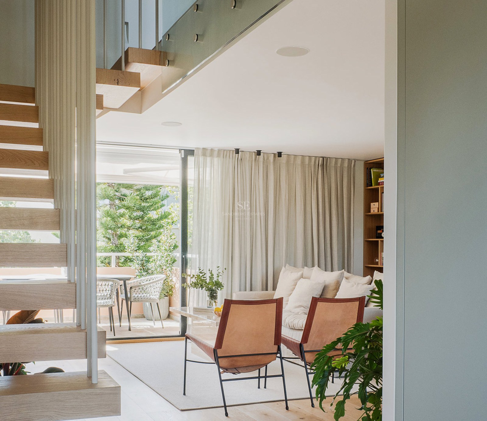 Vista de un salón luminoso con escaleras flotantes de madera, sillas de cuero y acceso a una terraza verde.