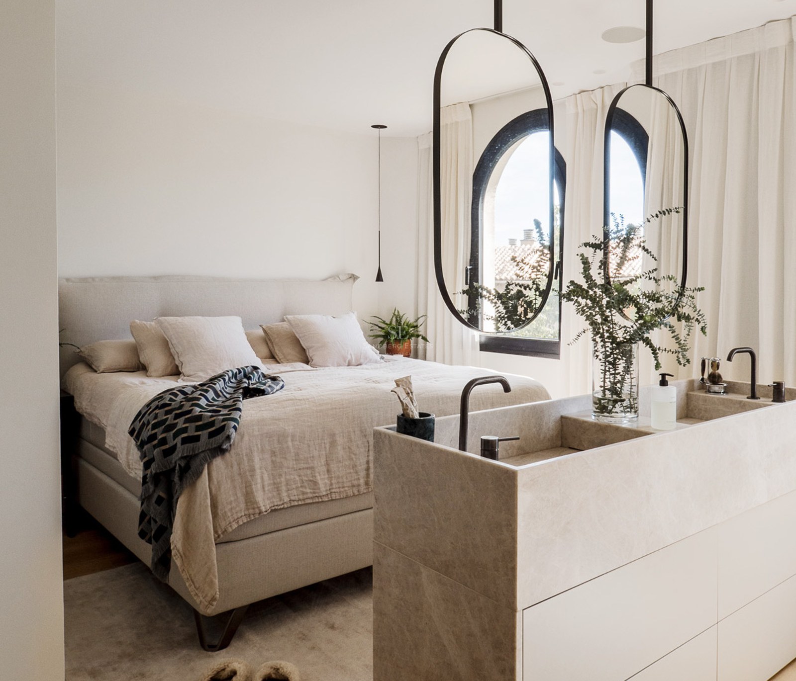 Dormitorio principal moderno con cama de tonos neutros y tocador de piedra integrado con espejos ovalados colgantes.
