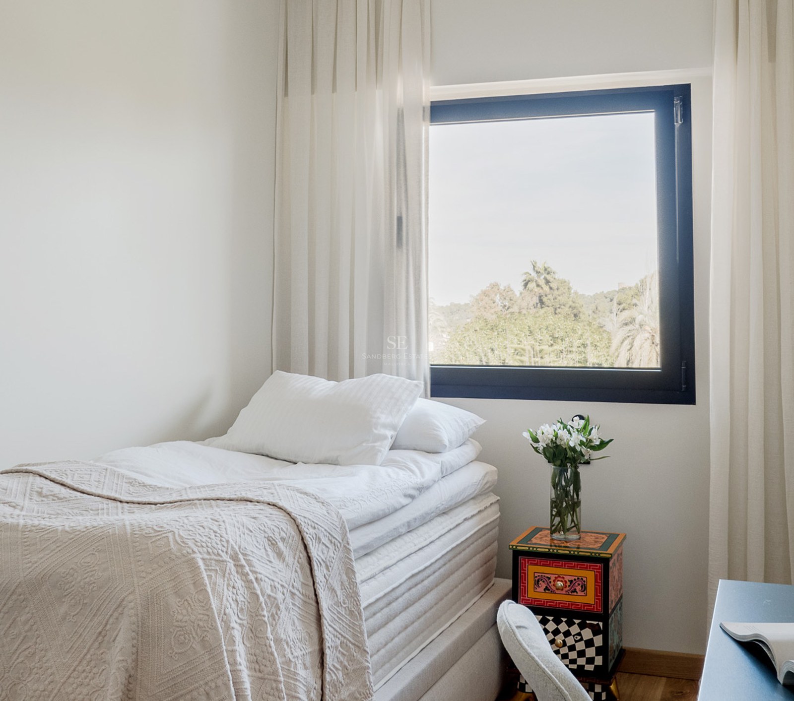 Cama individual con sábanas blancas junto a una ventana con vistas a árboles y una mesita de noche colorida.