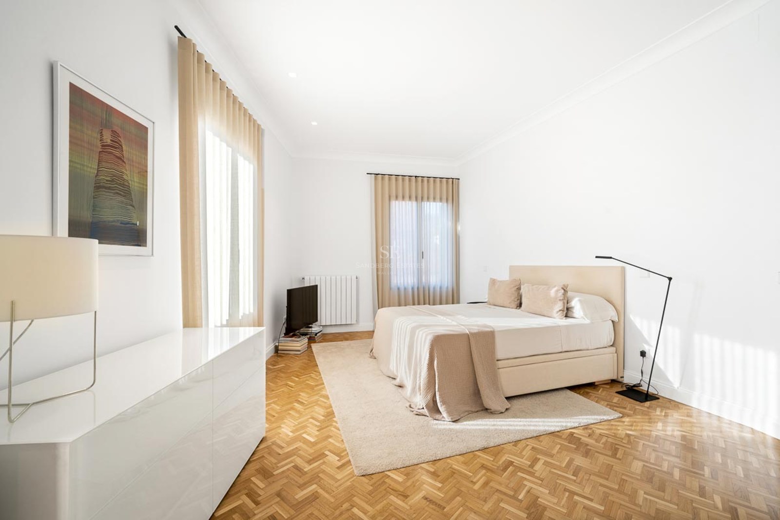 Chambre lumineuse avec parquet en chevrons, grand lit, rideaux beiges et lampe design.