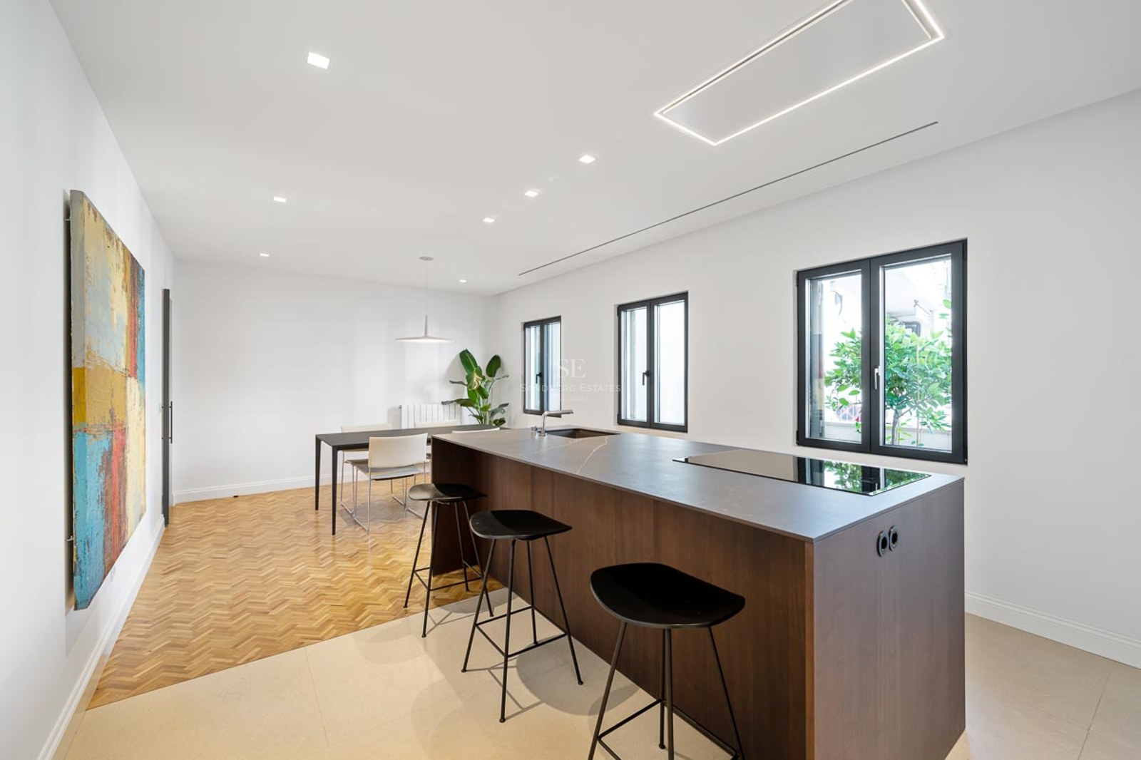 Cuisine minimaliste avec îlot central sombre, tabourets noirs, parquet à chevrons et tableau abstrait coloré.