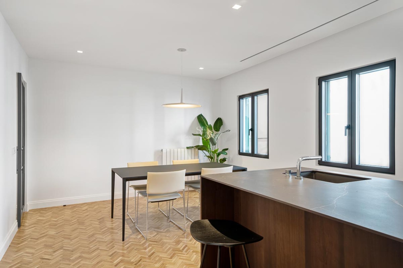 Salle à manger moderne avec table noire, chaises blanches, parquet en chevrons et îlot de cuisine.
