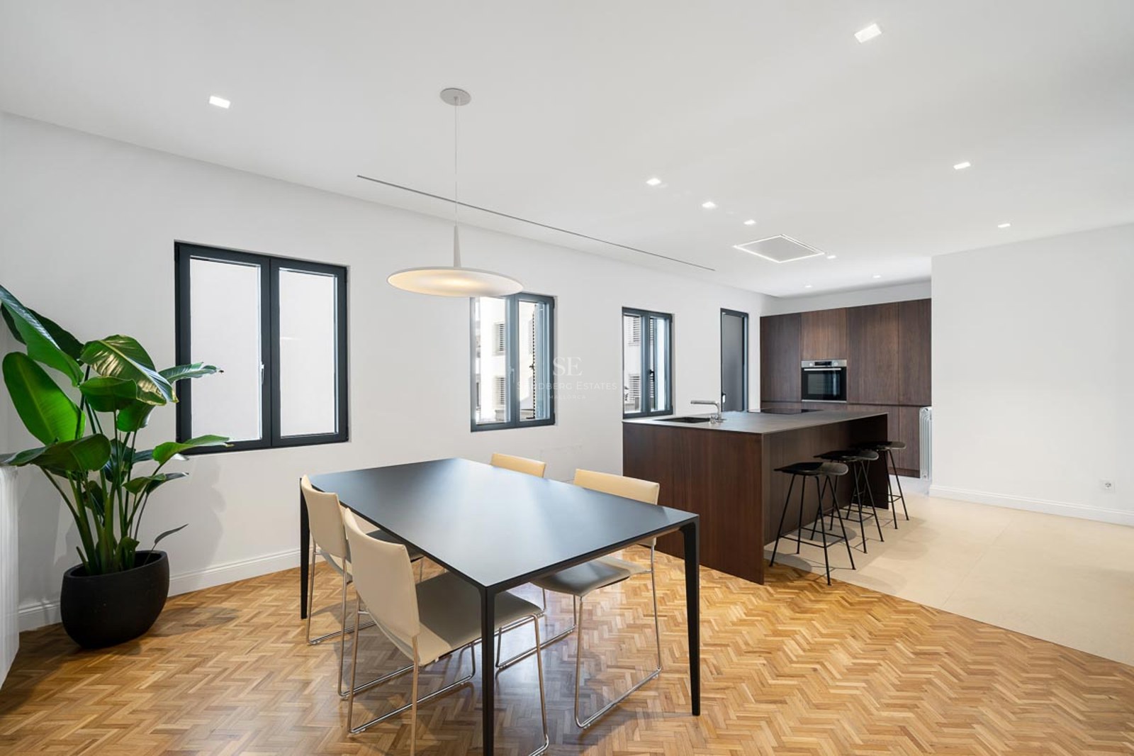 Cuisine moderne avec îlot en bois sombre et salle à manger avec parquet en chevrons et grandes fenêtres.