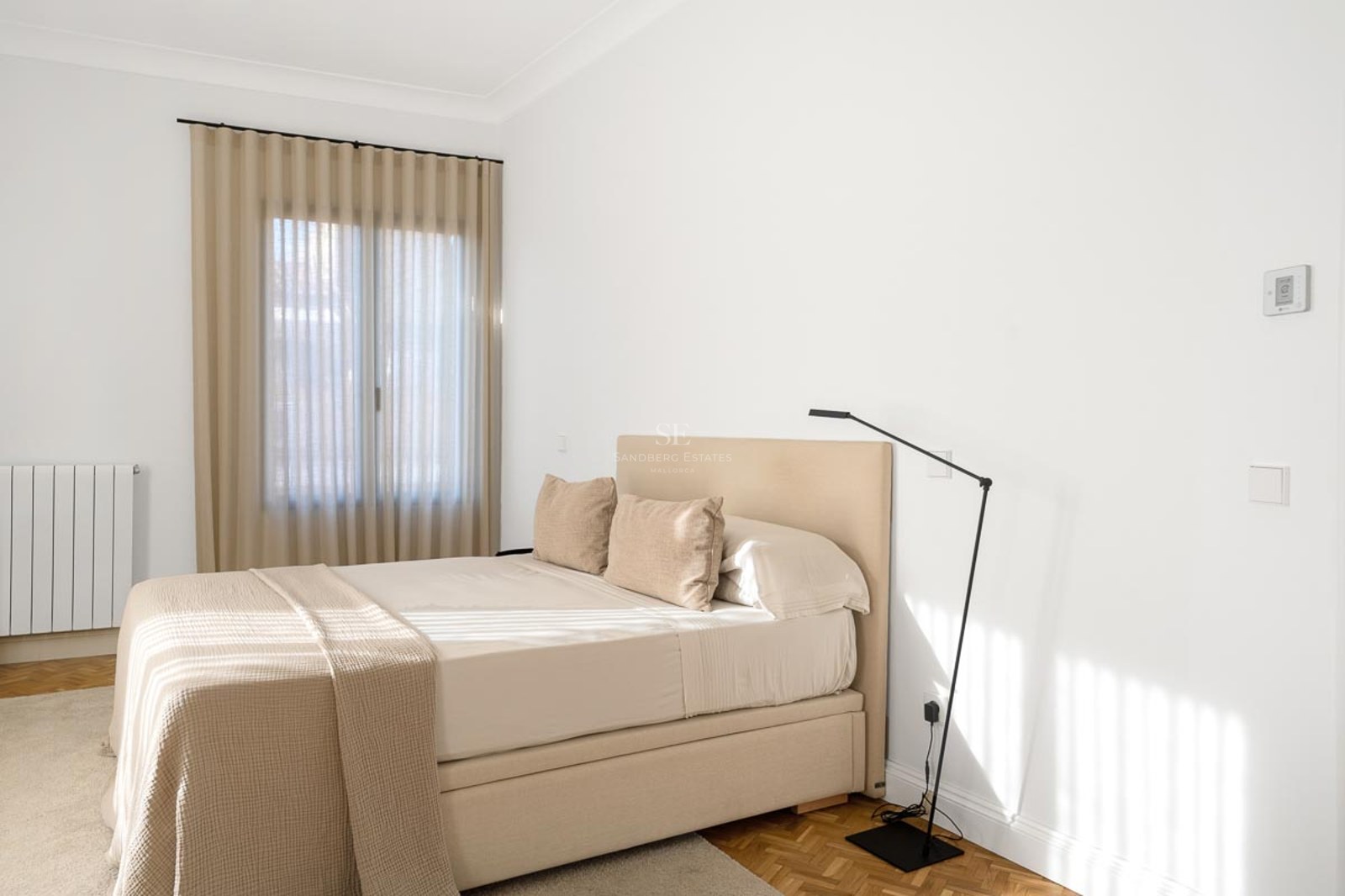 Chambre lumineuse avec un lit beige, des murs blancs et un parquet en bois chaleureux.
