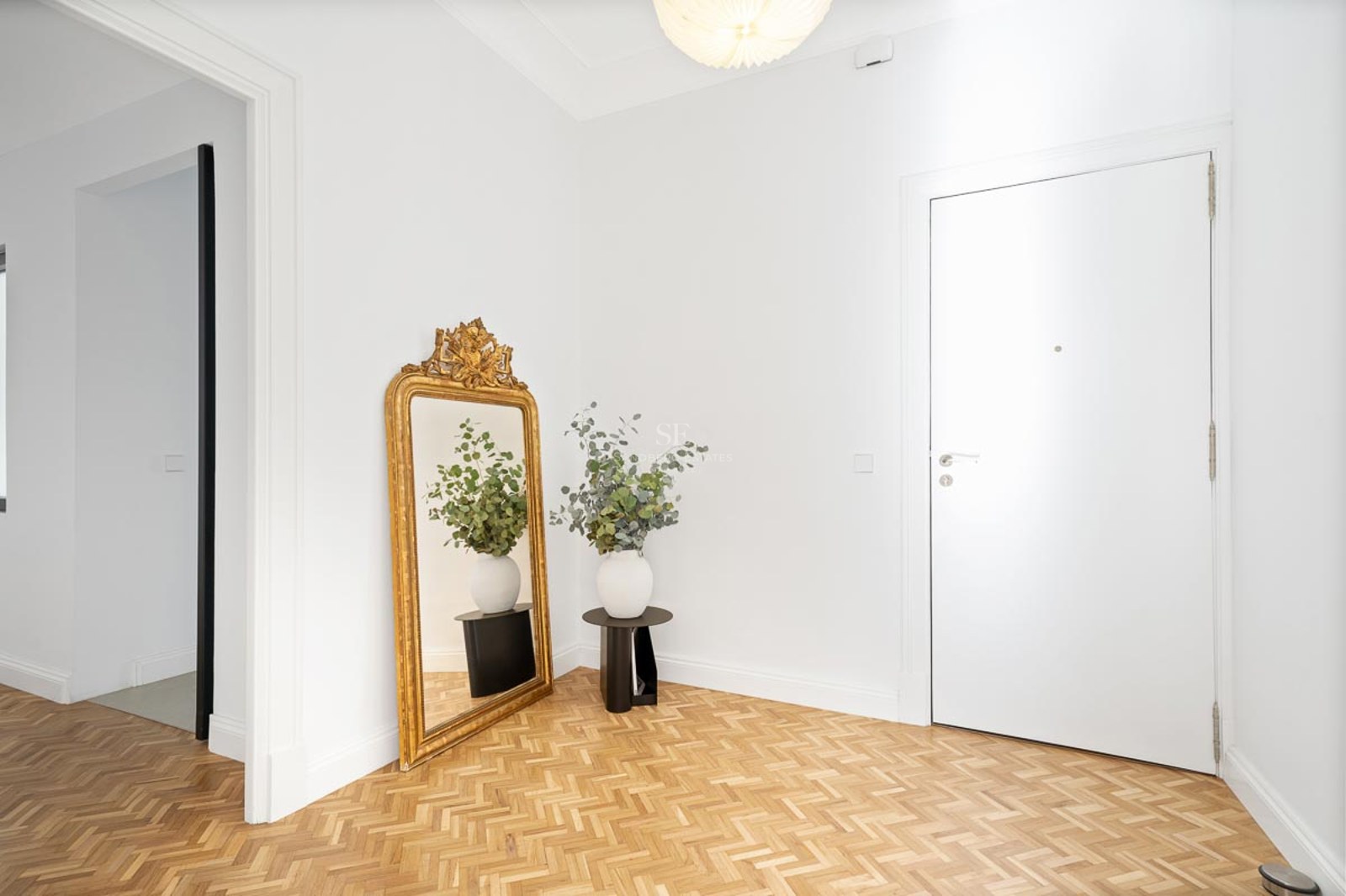 Entrée lumineuse avec murs blancs, parquet en chevrons, un grand miroir doré et une porte blanche.