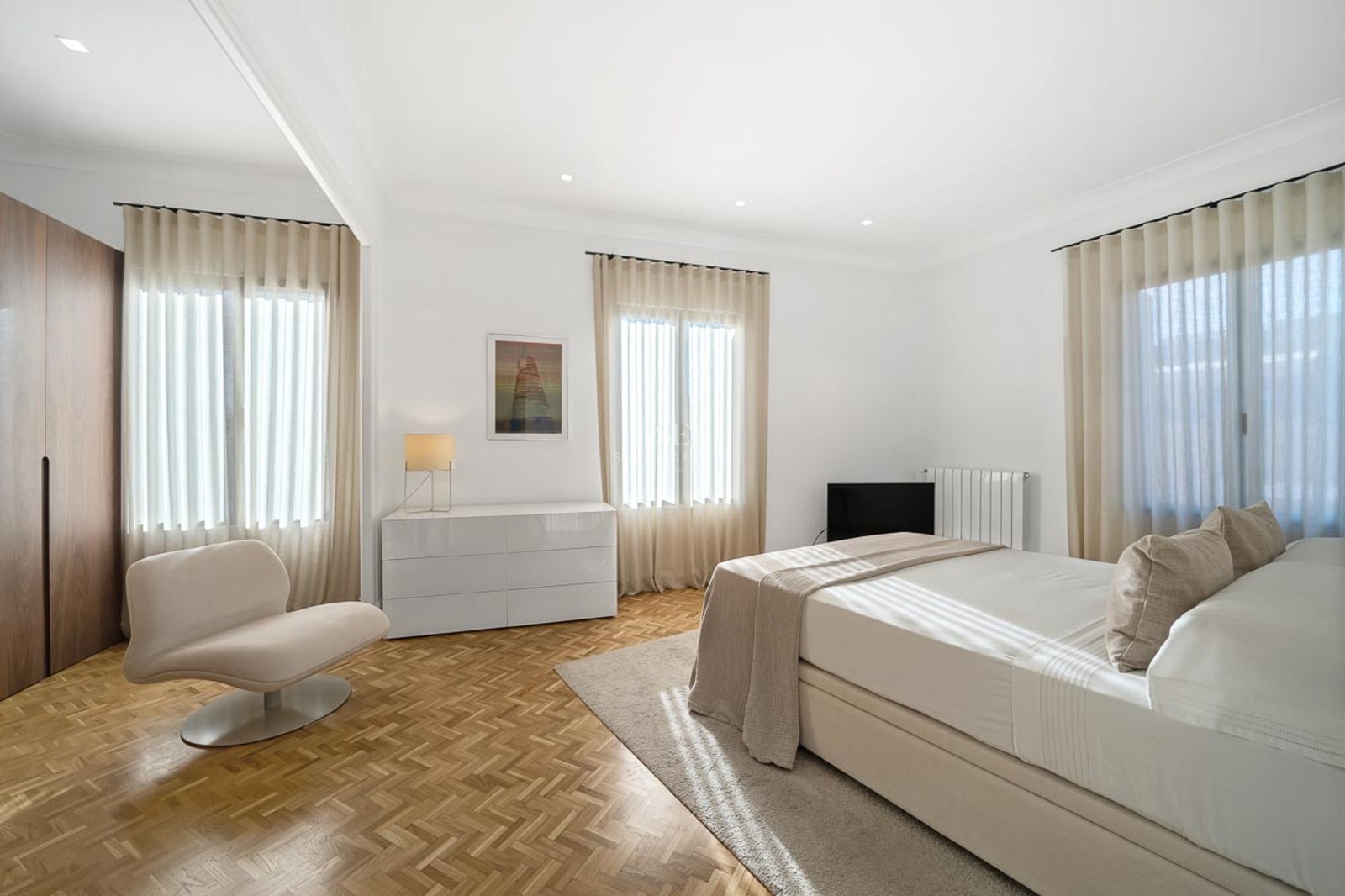 Chambre lumineuse avec parquet en chevrons, lit double, fauteuil design et rideaux voilages.