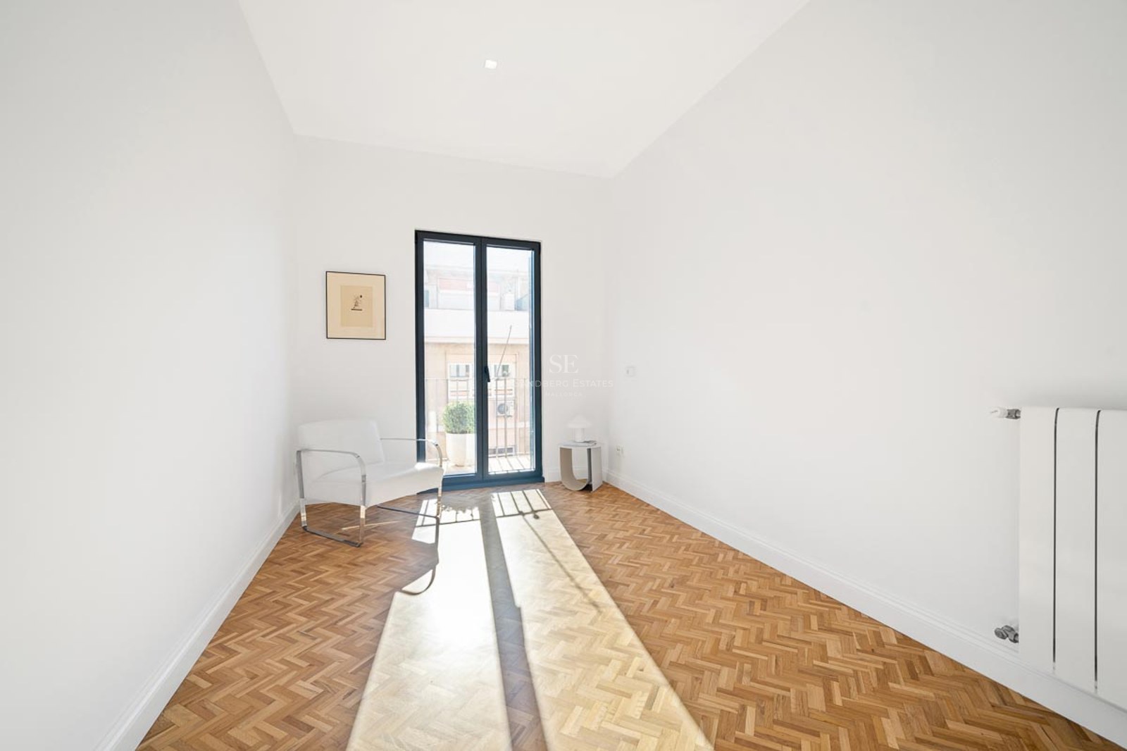 Pièce blanche lumineuse avec parquet en bois en chevrons, fauteuil blanc et porte-fenêtre donnant sur un balcon.