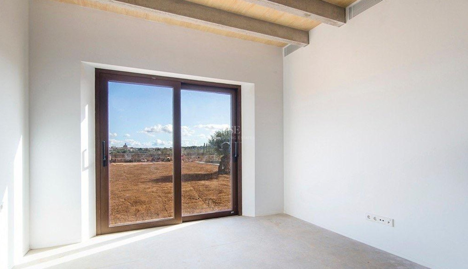 Empty modern room with white walls, wooden ceiling beams, and a sliding glass door overlooking a Mediterranean landscape.