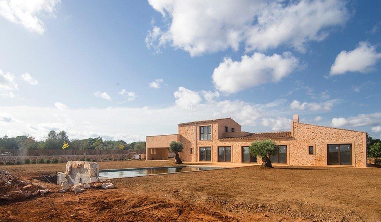 Natural stone facade of a modern villa with a swimming pool and olive trees under a bright blue sky.