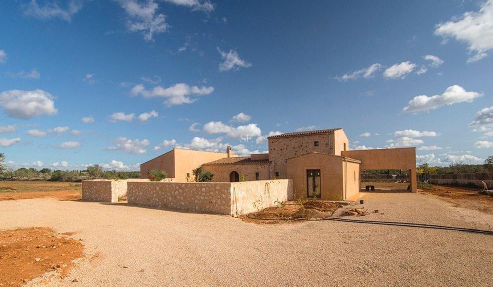 Exterior view of a modern Mediterranean stone villa with ochre walls, gravel driveway, and clear blue sky.